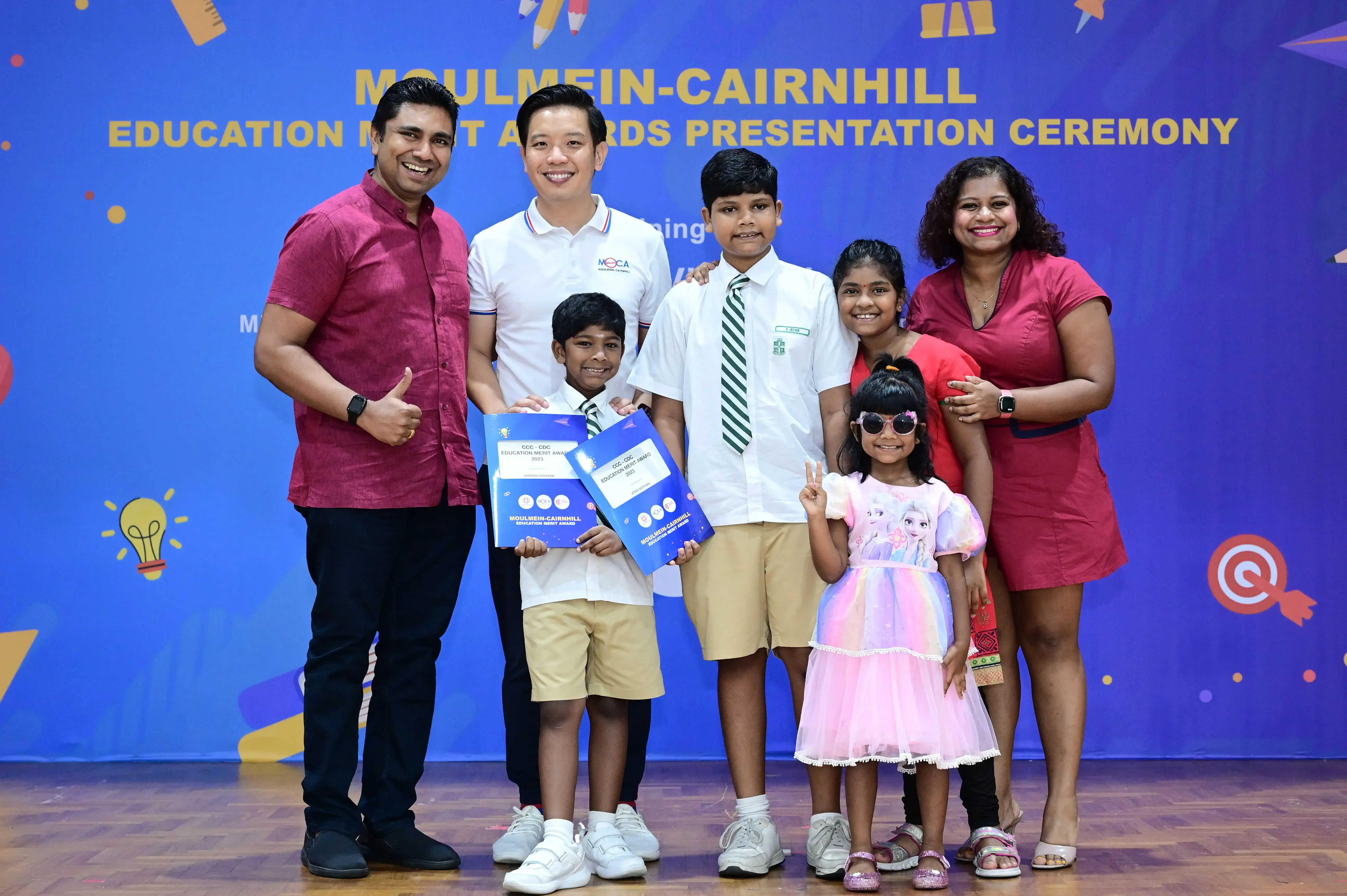 Group of adults and children posing at Moulmein-Cairnhill Education Merit Awards Presentation Ceremony, children holding award folders.