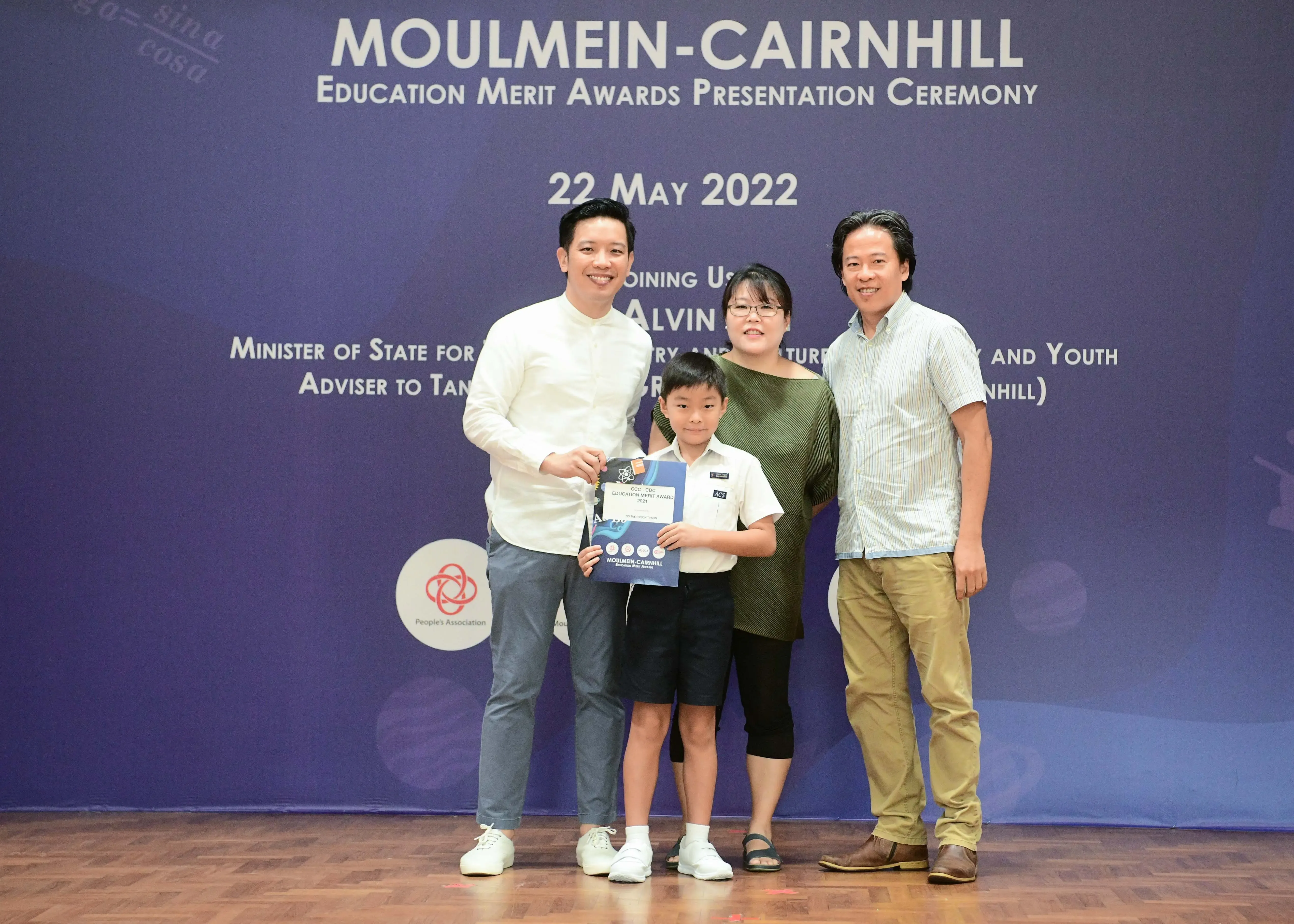 Four people pose before a Moulmein-Cairnhill Education Merit Awards banner dated 22 May 2022, with a boy holding a certificate.