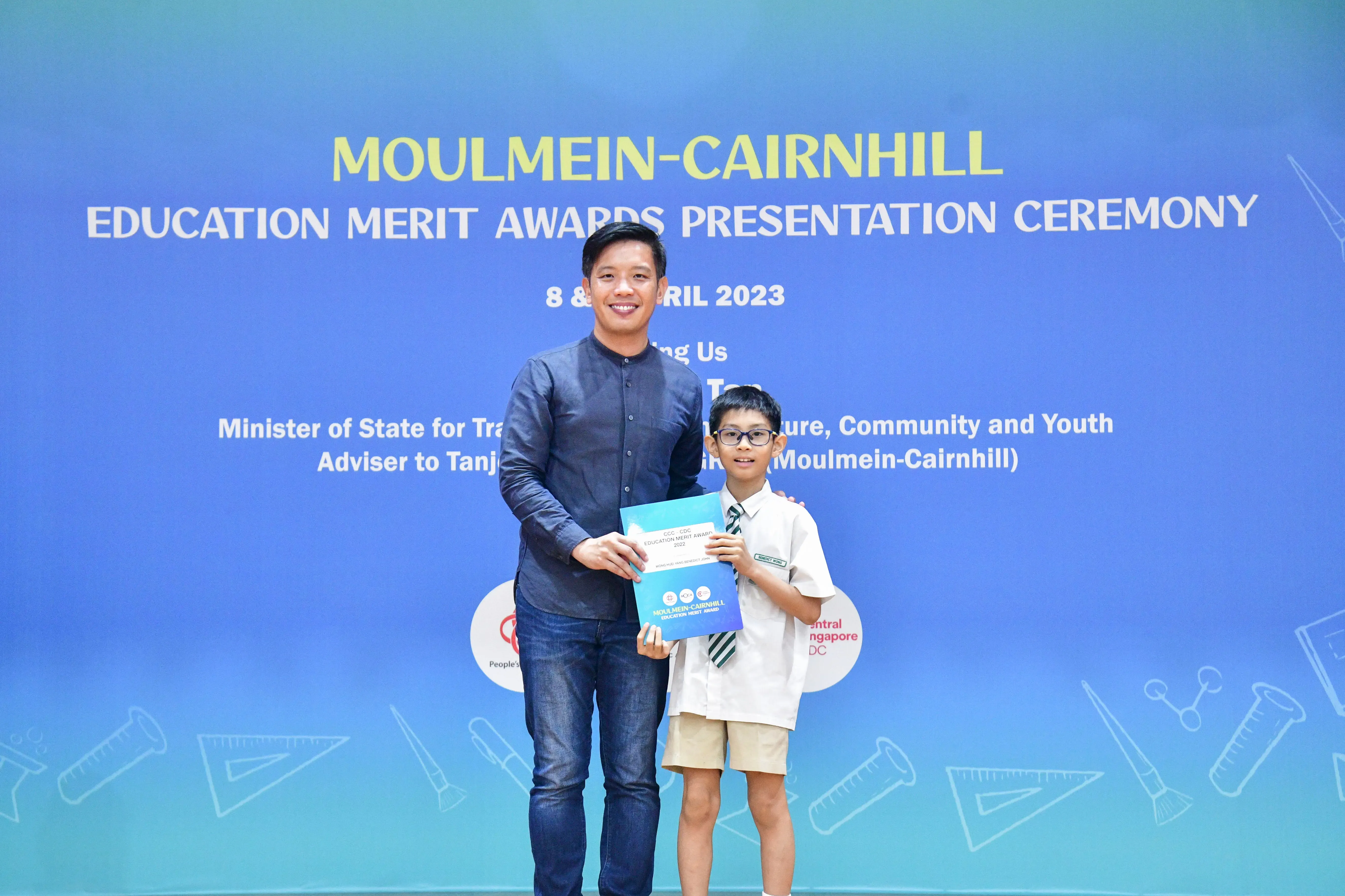 A man and a young boy hold a certificate together in front of a Moulmein-Cairnhill Education Merit Awards banner.