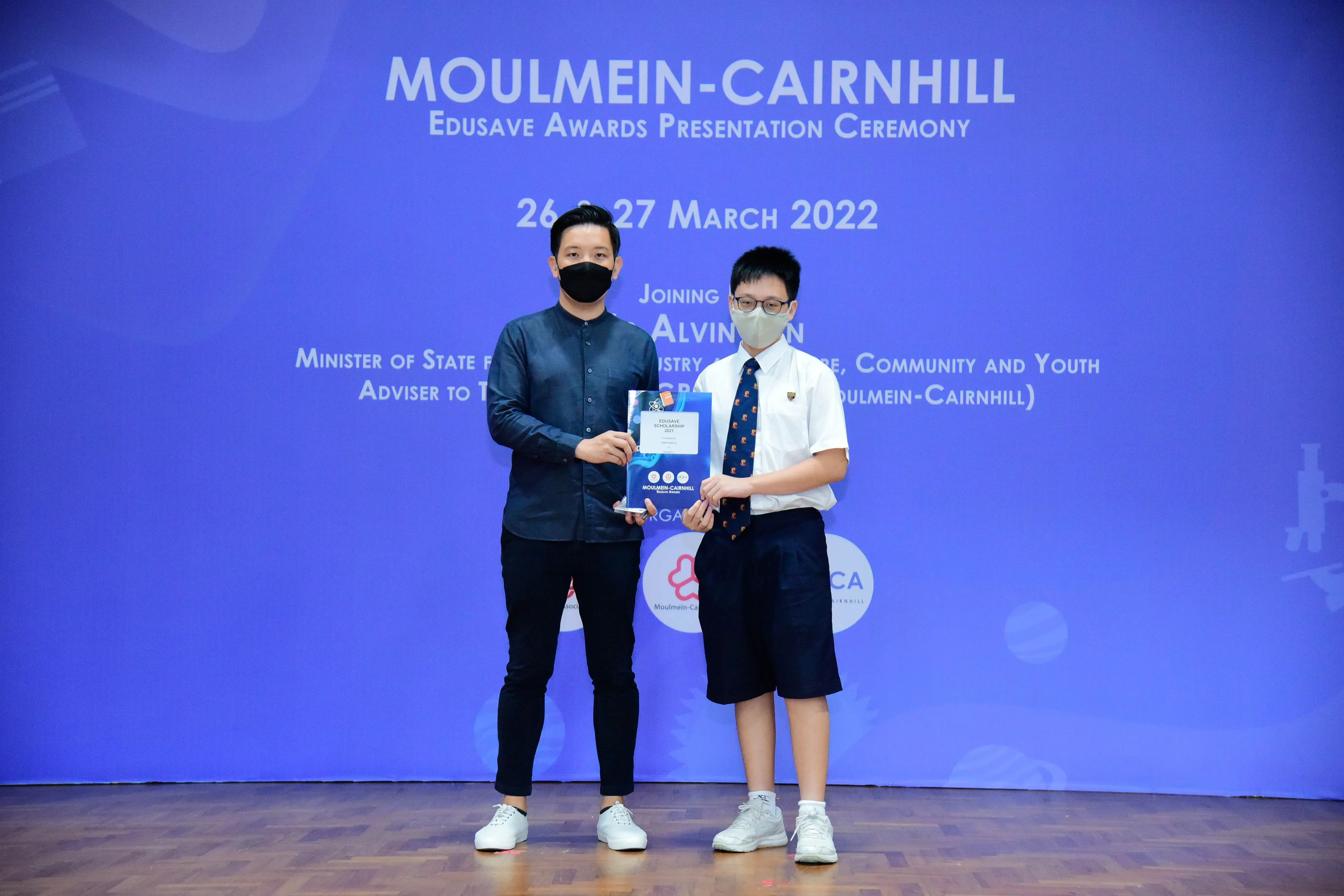 Two masked individuals hold an award at the Moulmein-Cairnhill Edusave Awards Presentation Ceremony, March 2022.