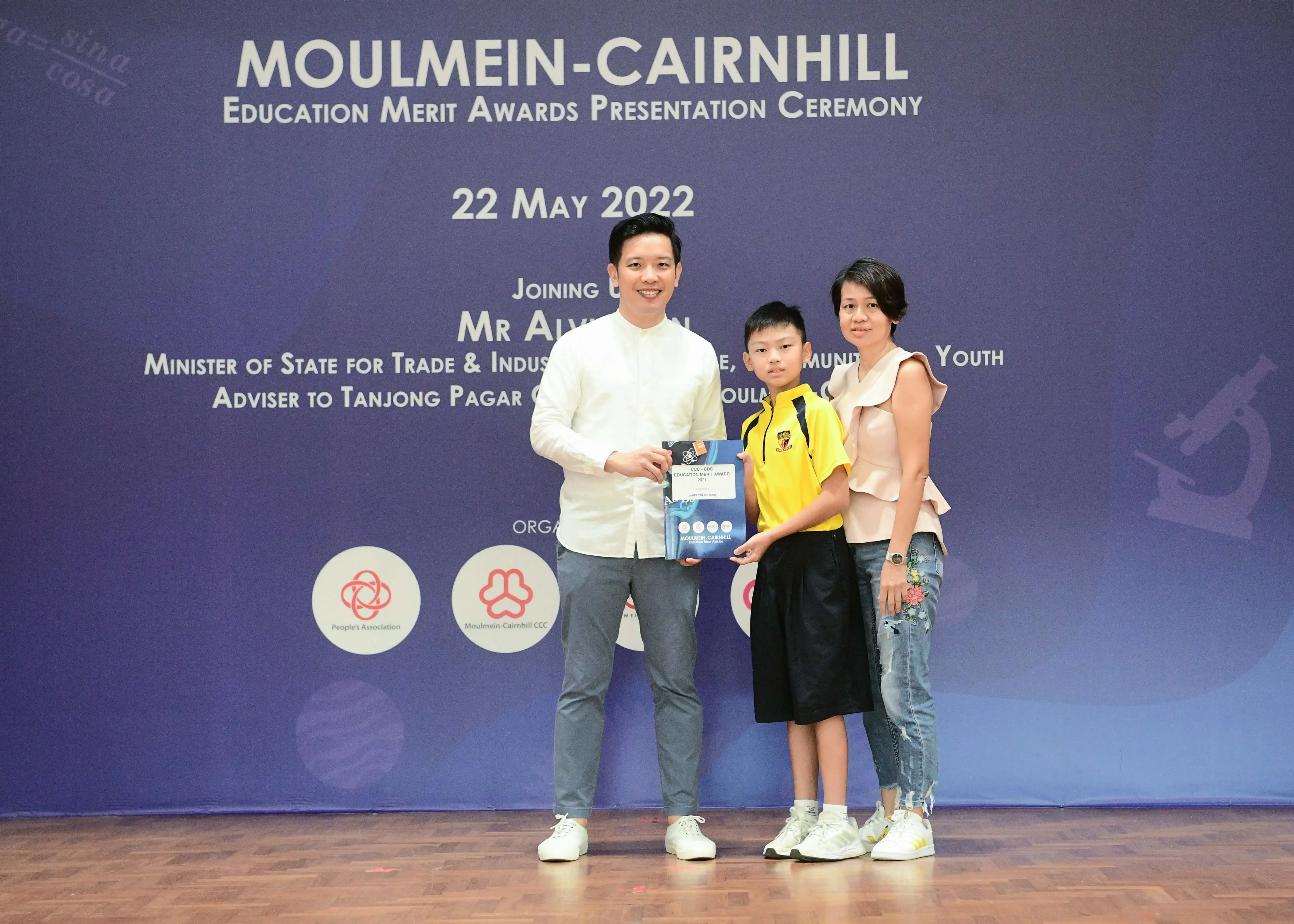 Two adults and a child hold an award at the Moulmein-Cairnhill Education Merit Awards Ceremony on 22 May 2022.