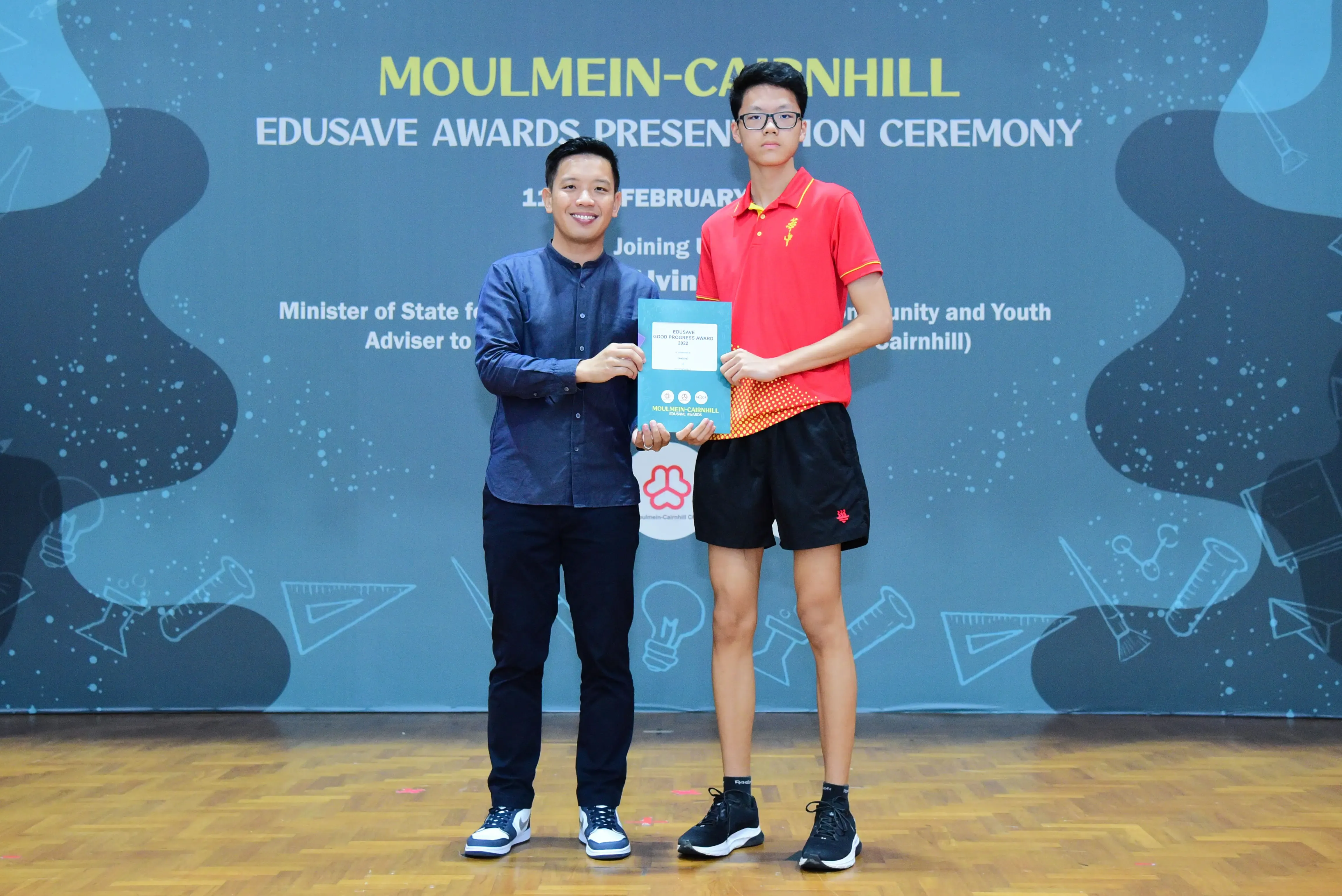 Two men hold a certificate together at the Moulmein-Cairnhill Edusave Awards Presentation Ceremony backdrop.