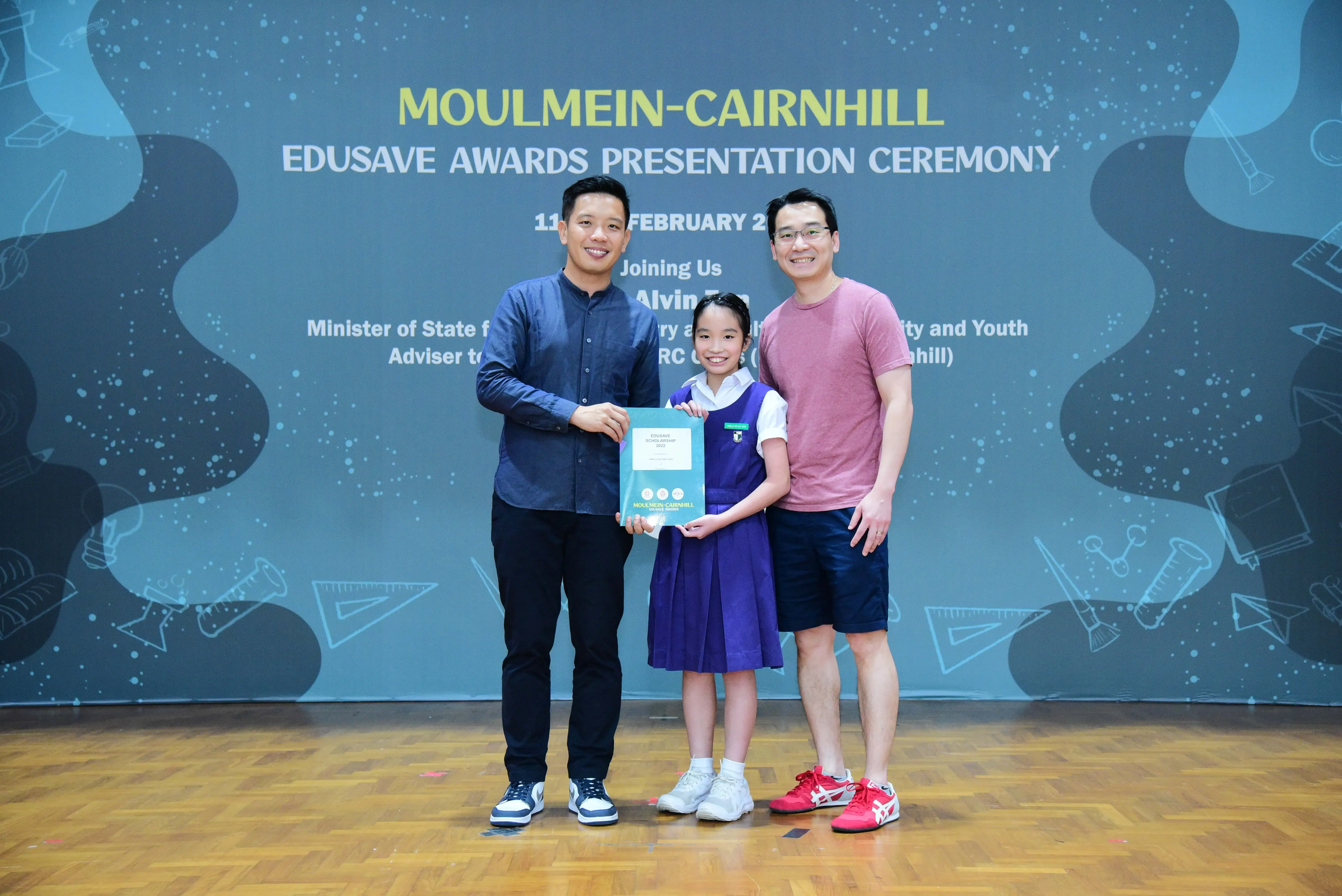 Two men and a young girl in school uniform hold a certificate at the Moulmein-Cairnhill Edusave Awards Presentation Ceremony.