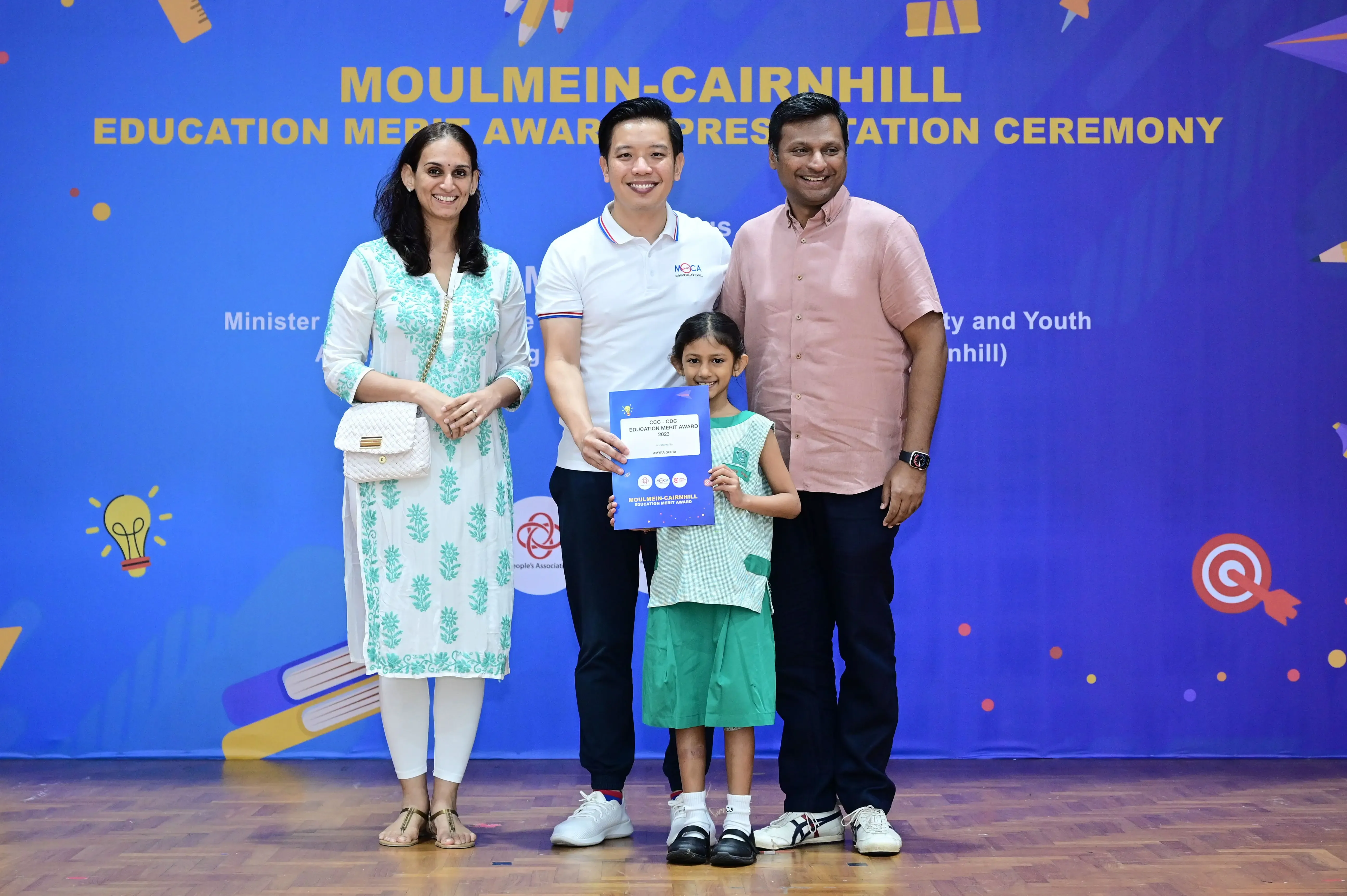 Four people pose at the Moulmein-Cairnhill Education Merit Award Presentation Ceremony, with a child holding a certificate.