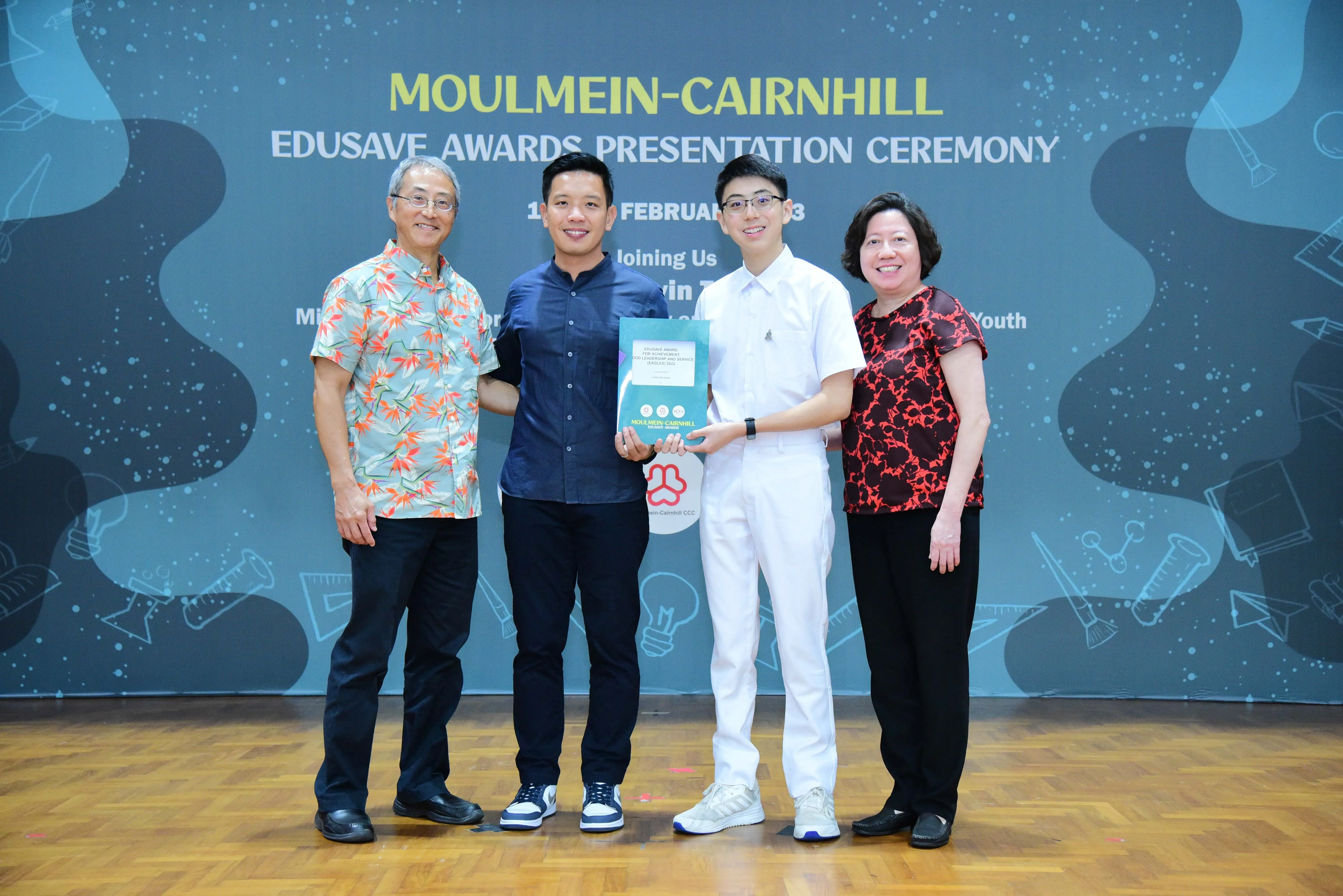 Four people stand smiling before a Moulmein-Cairnhill Edusave Awards Presentation Ceremony banner, holding an award booklet.
