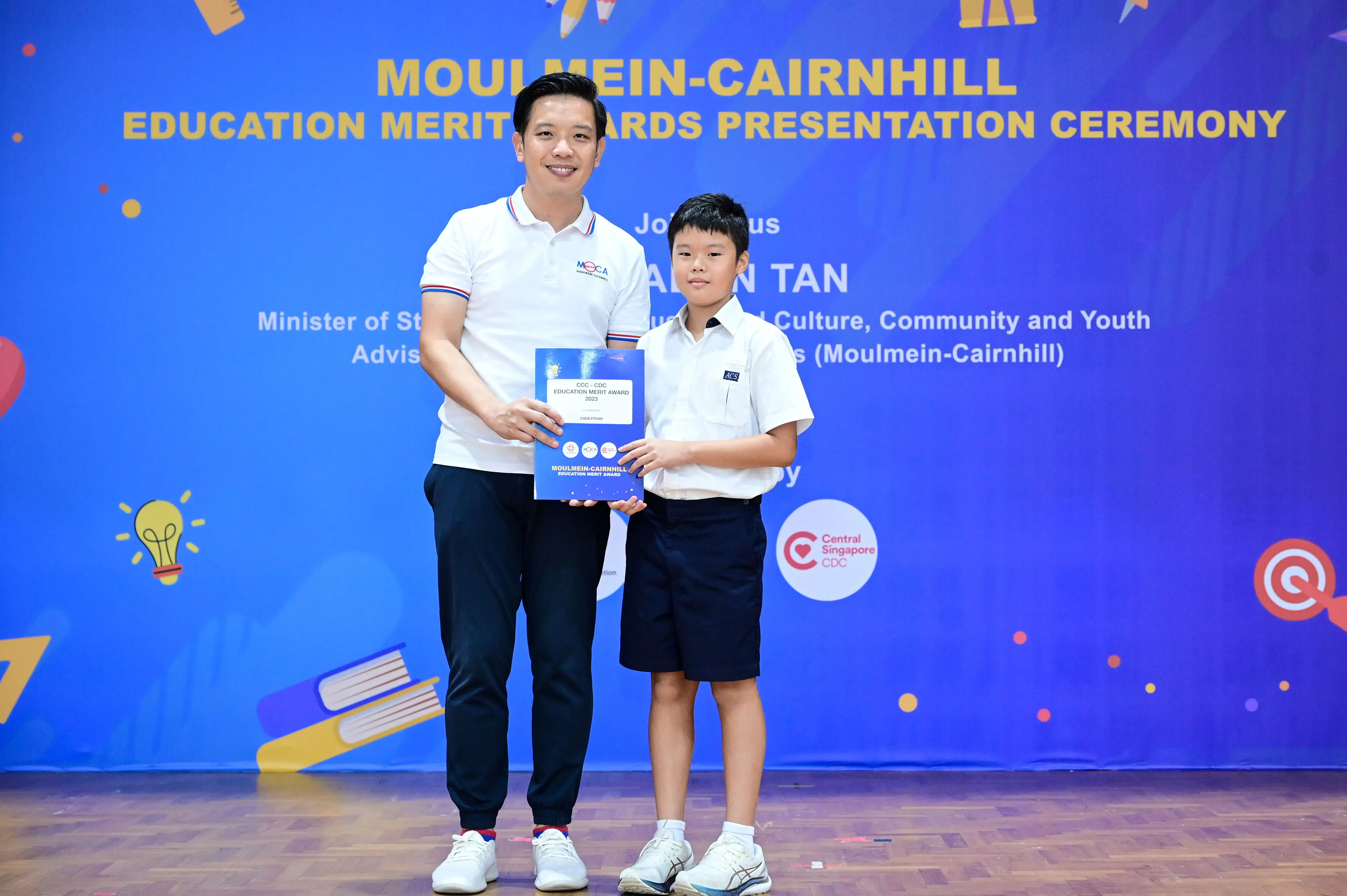 An adult and a young boy hold a certificate together on stage at the Moulmein-Cairnhill Education Merit Awards Ceremony.