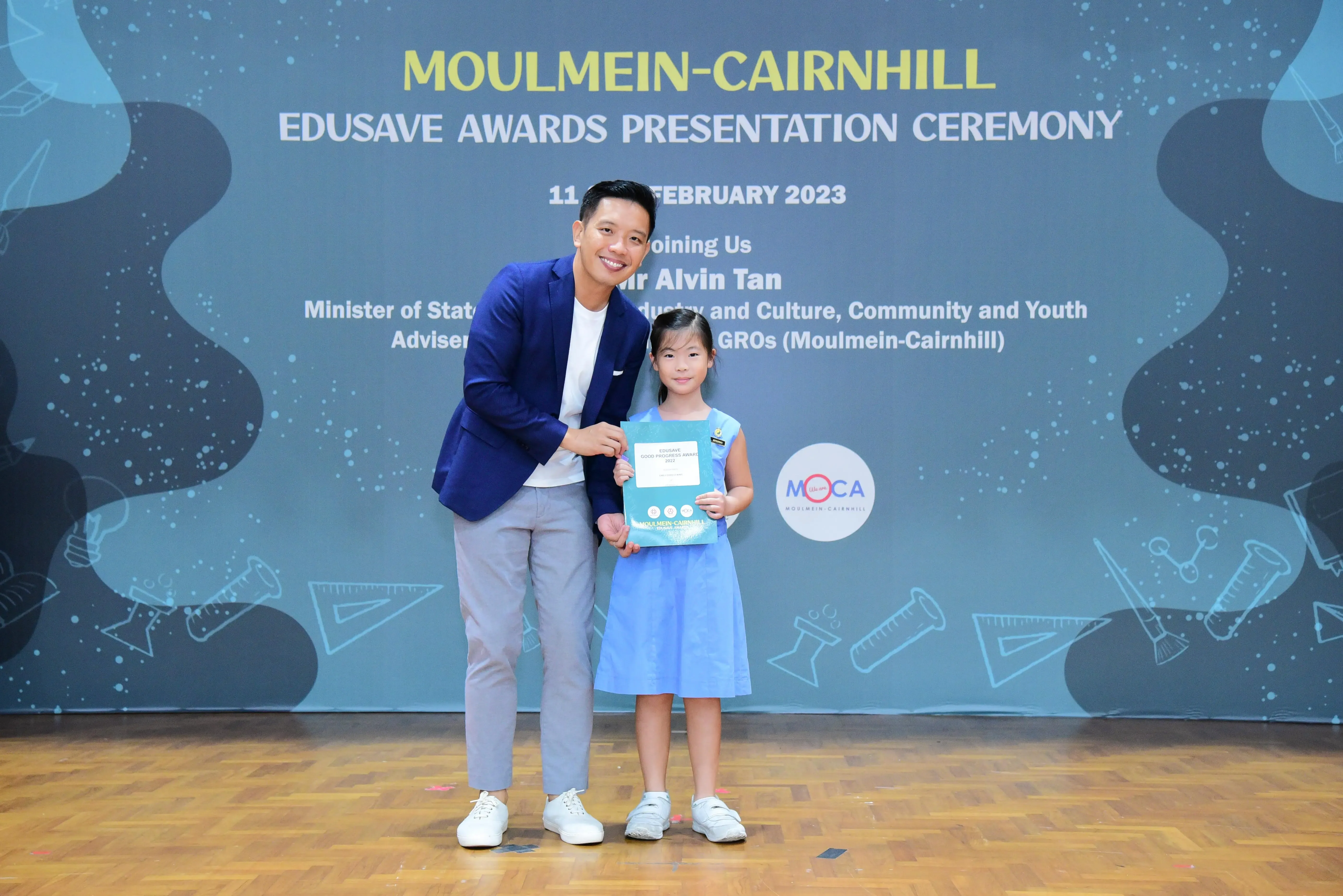 A man in a blue blazer and a young girl in a blue dress hold a certificate at the Moulmein-Cairnhill Edusave Awards 2023.