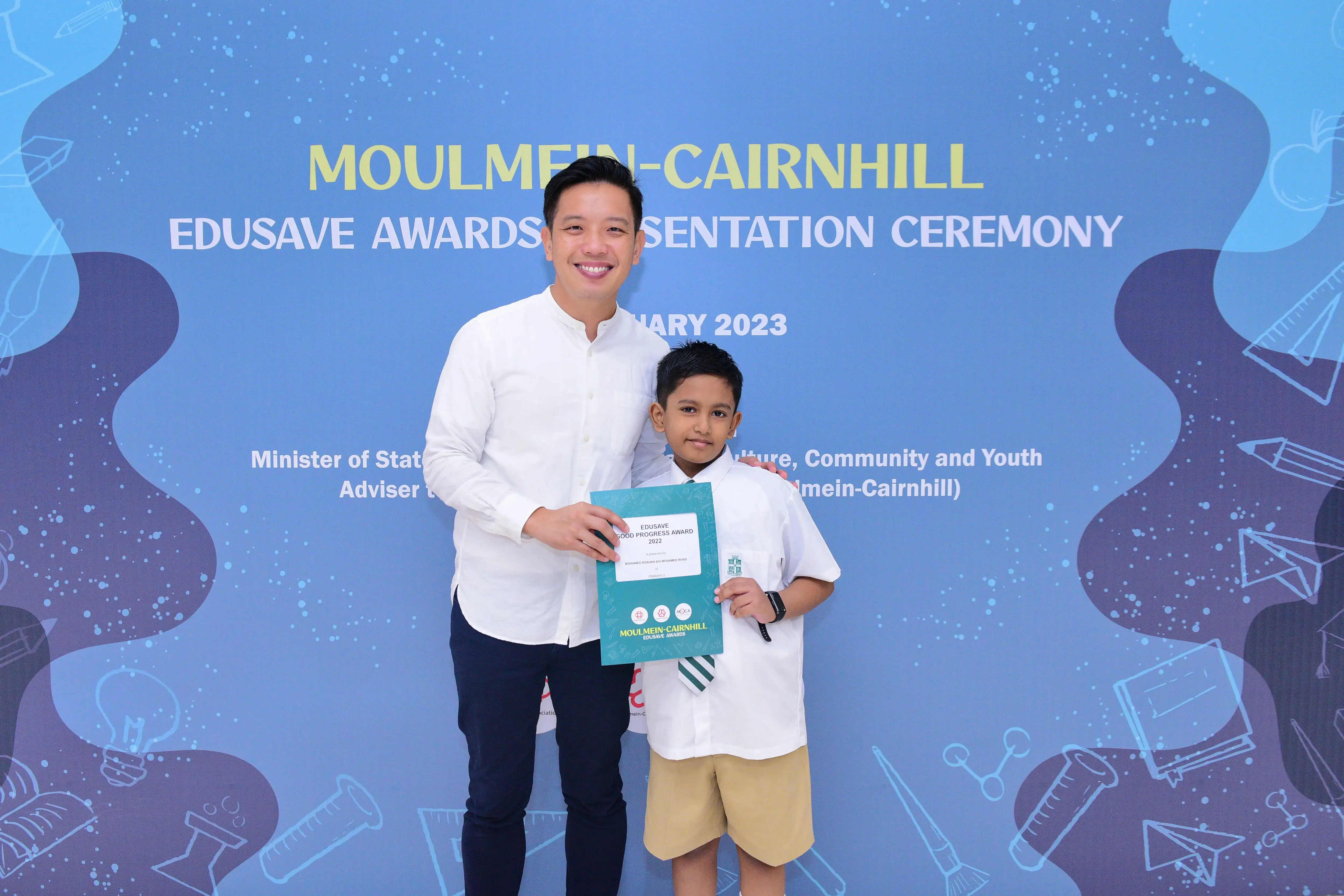 A man and a young boy hold a teal award folder at the Moulmein-Cairnhill Edusave Awards Ceremony 2023.