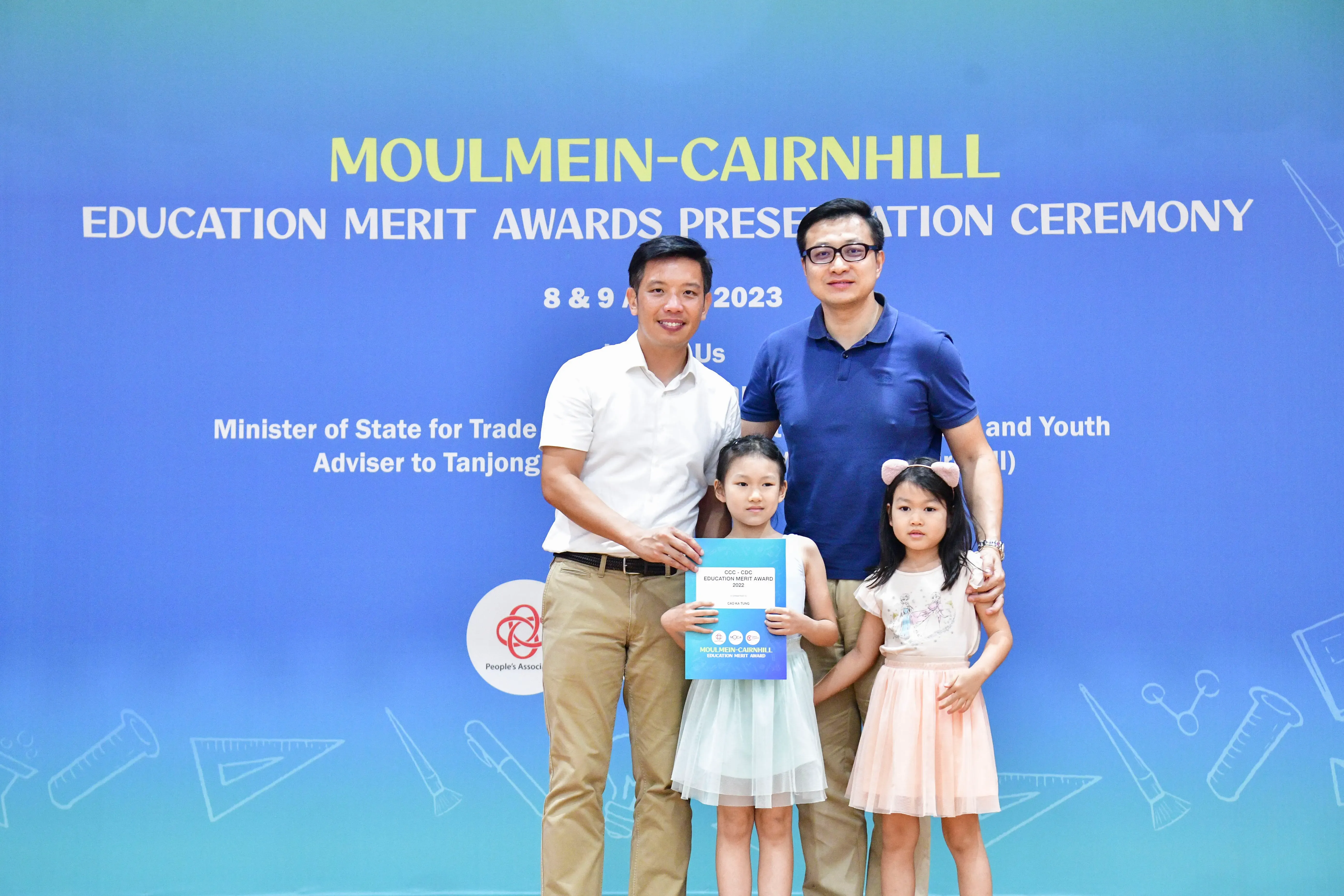 Two men and two young girls pose at the Moulmein-Cairnhill Education Merit Awards Ceremony 2023, one girl holding a certificate.
