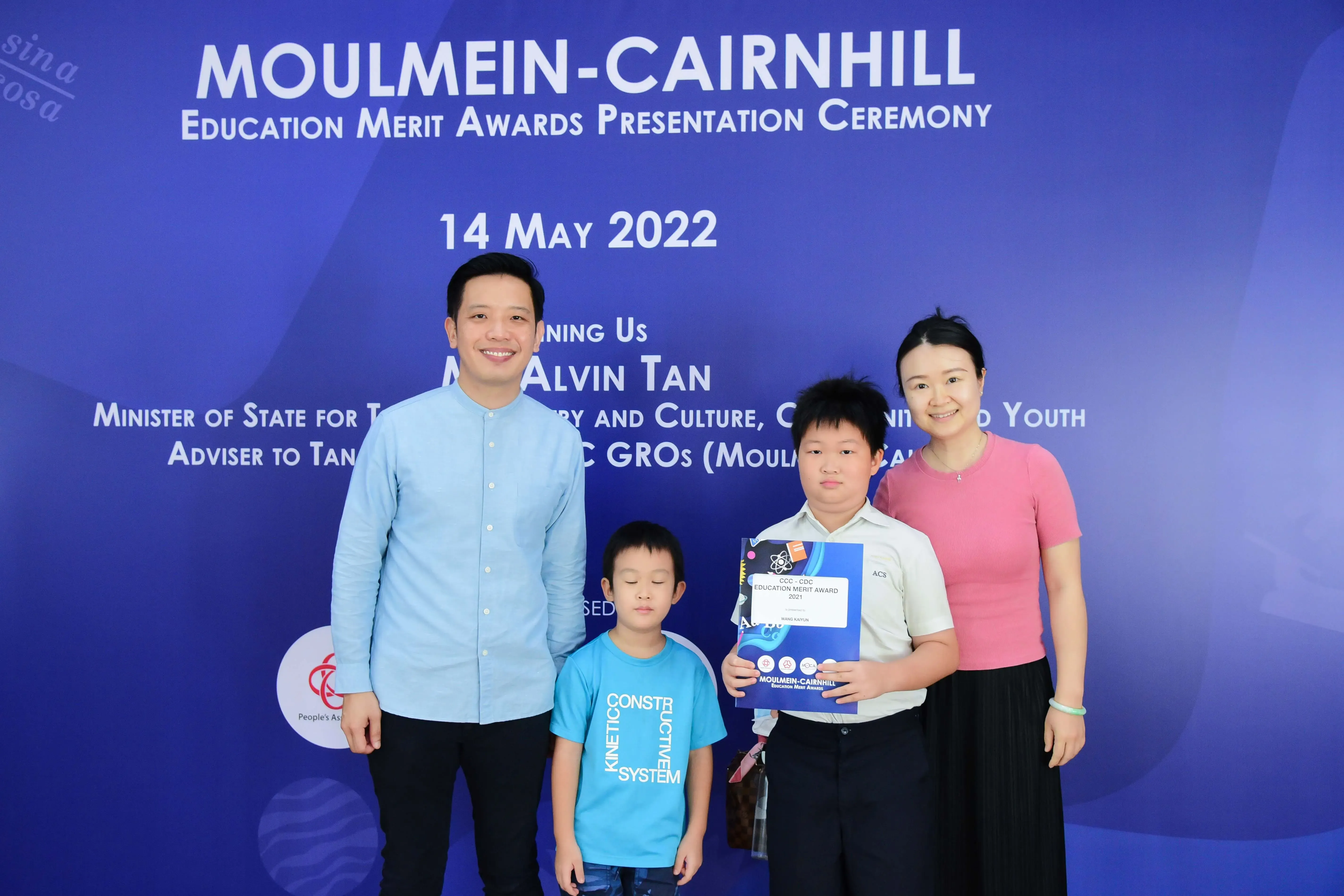 Four people pose before a blue Moulmein-Cairnhill Education Merit Awards banner dated 14 May 2022.
