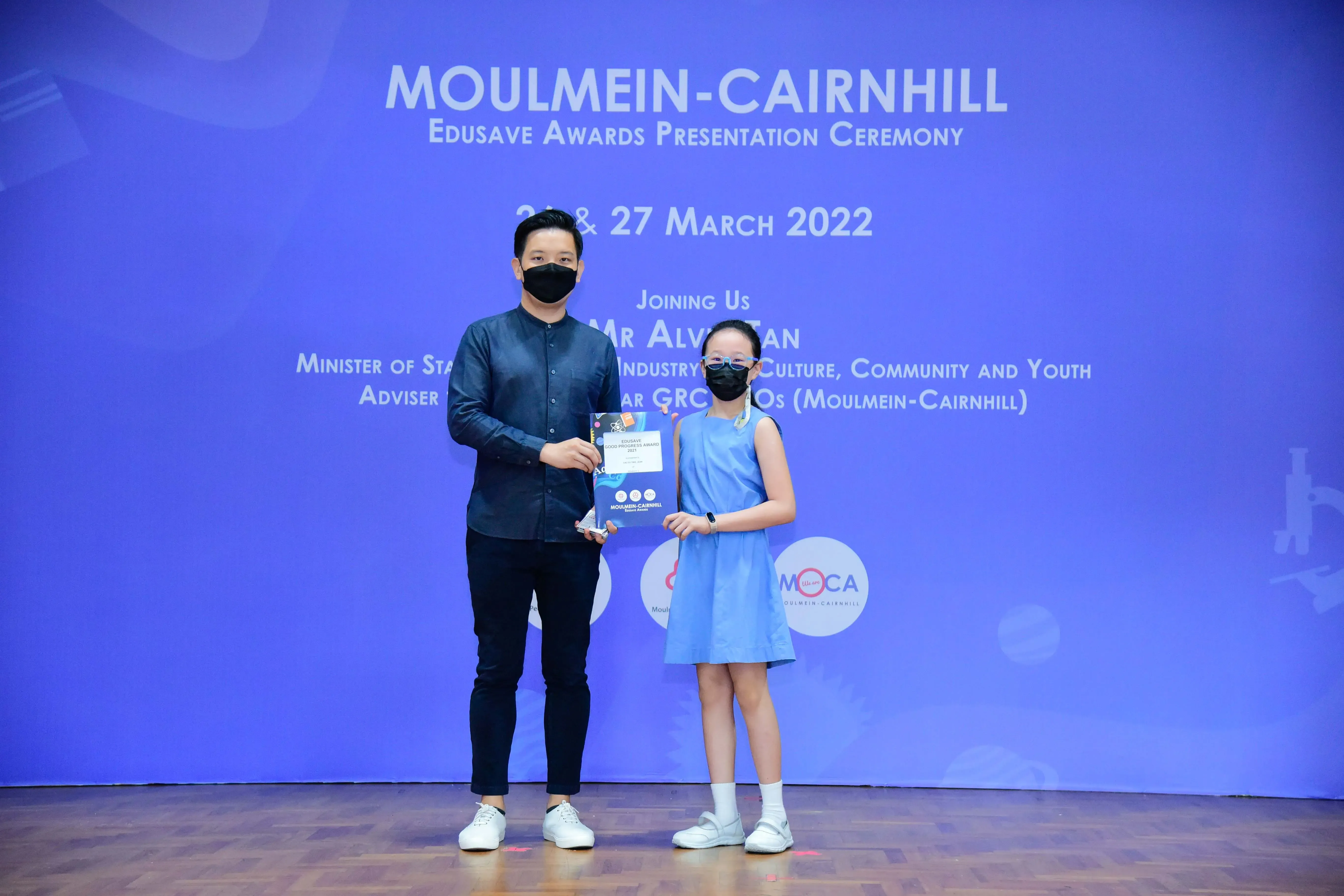 An adult and a young girl wearing masks pose with an award at the Moulmein-Cairnhill Edusave Awards Ceremony, March 2022.