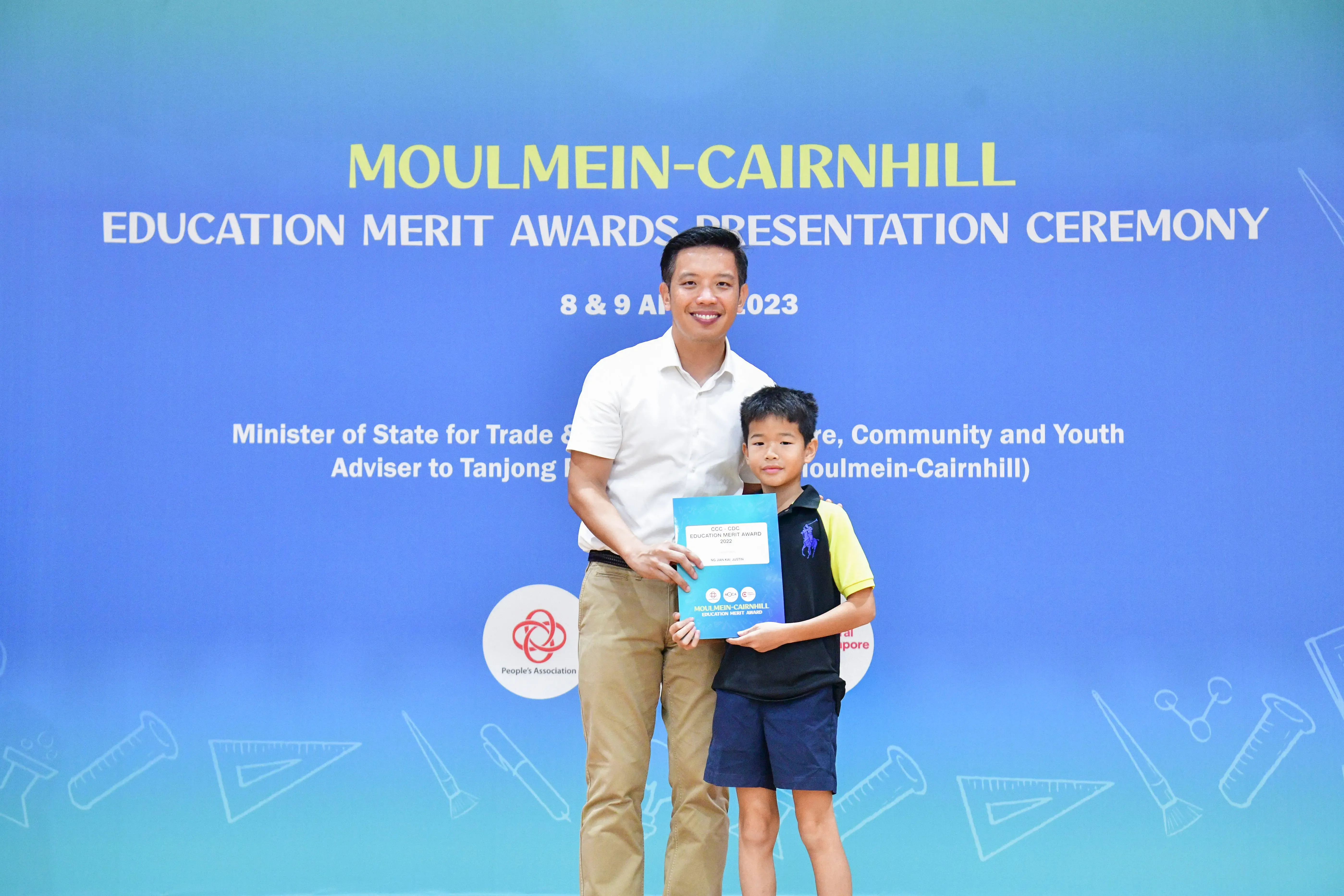 A man and a young boy hold an award certificate in front of a Moulmein-Cairnhill Education Merit Awards banner.