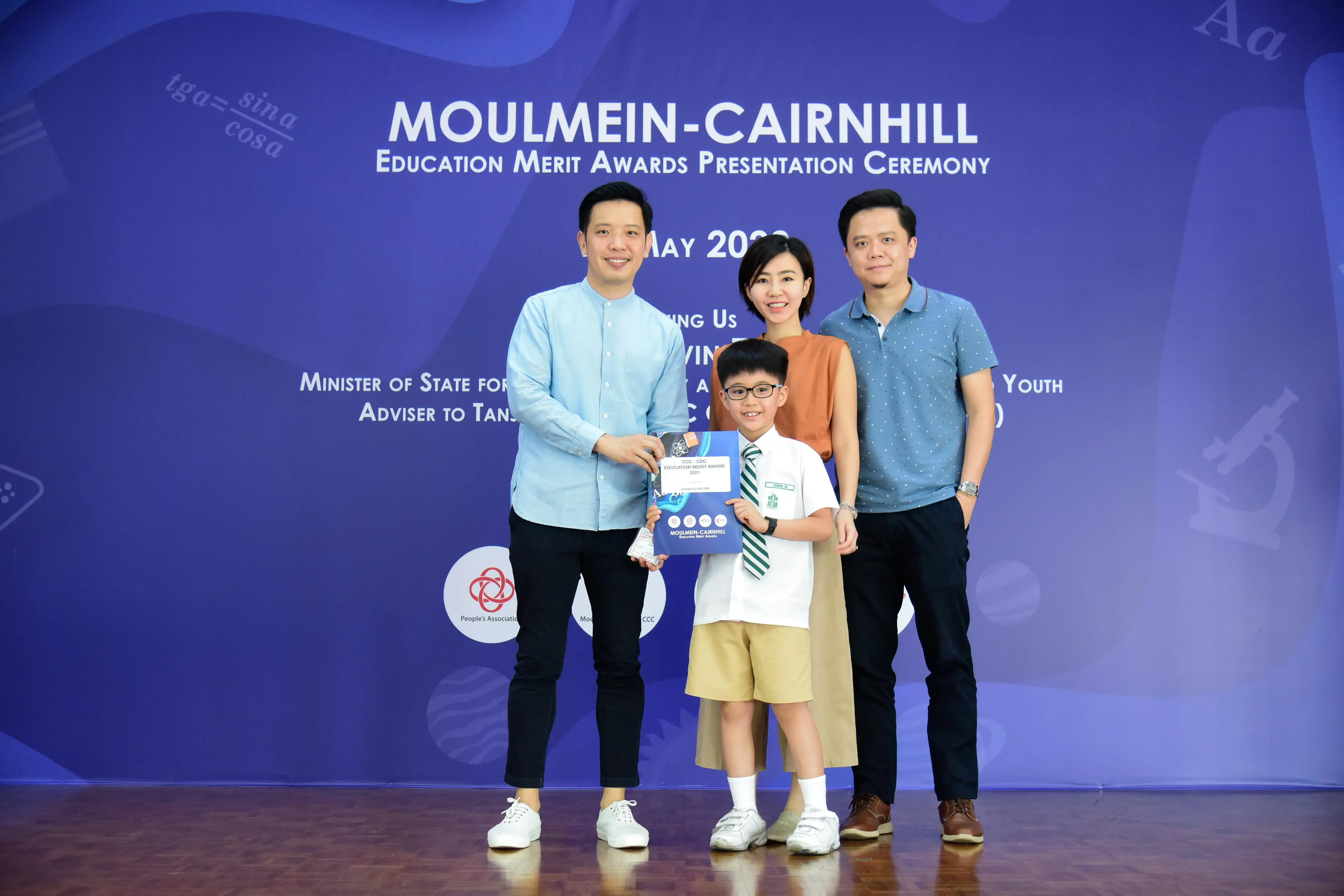 Three adults and a uniformed boy holding an award certificate at the Moulmein-Cairnhill Education Merit Awards ceremony.