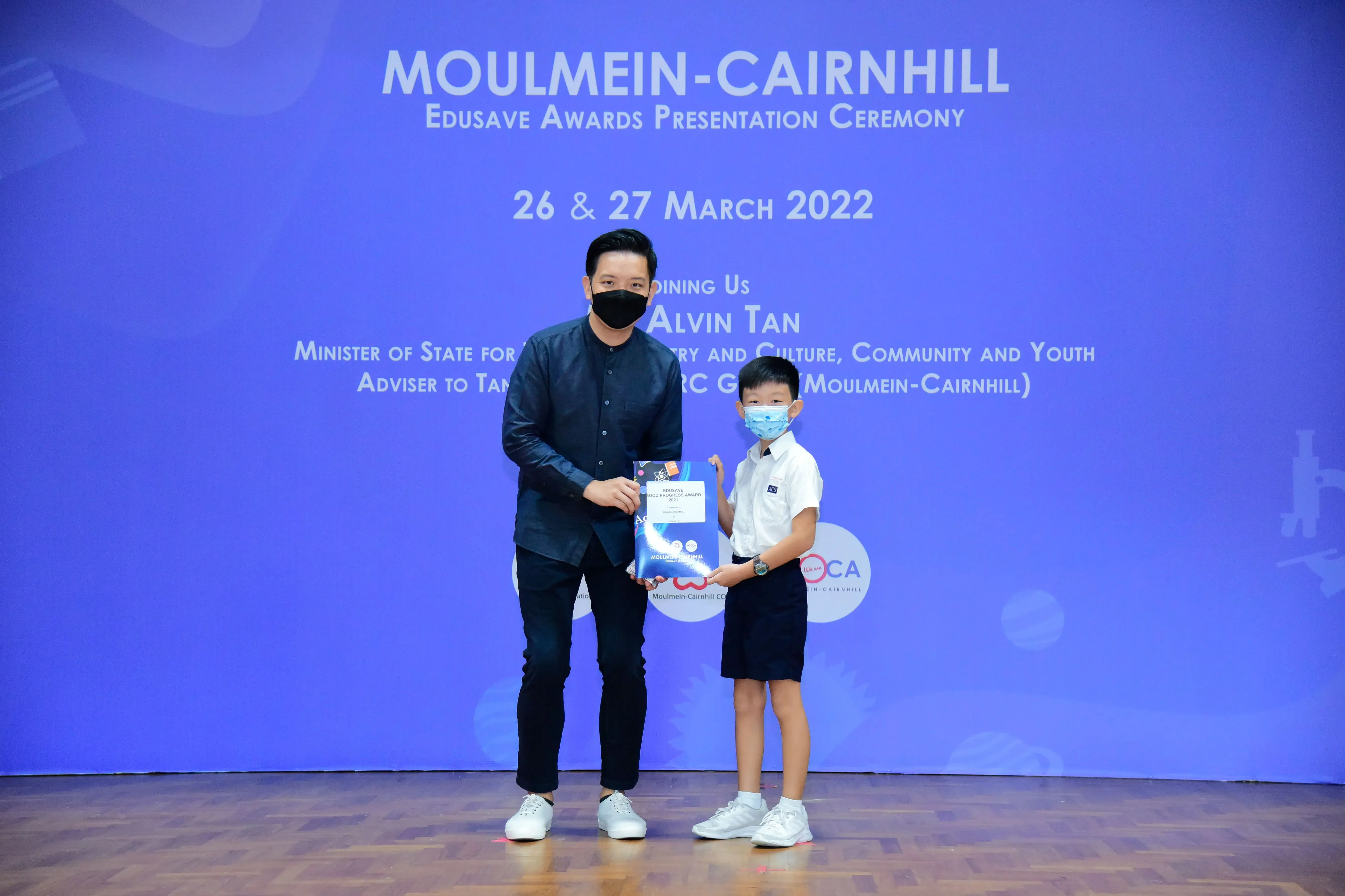 An adult and a young boy wearing masks hold an award at the Moulmein-Cairnhill Edusave Awards Ceremony 2022.