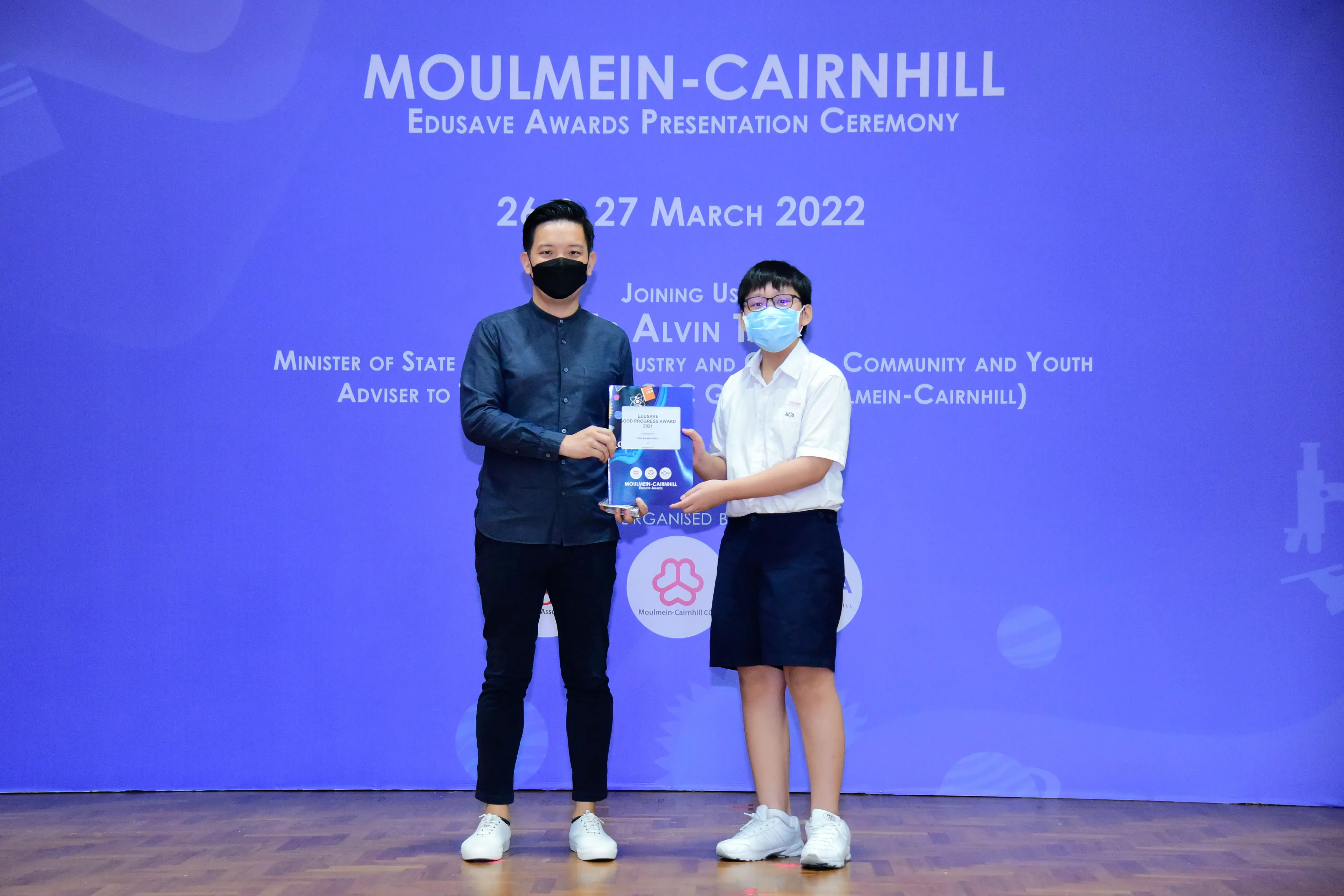 Two masked individuals hold an award together on stage at the Moulmein-Cairnhill Edusave Awards Ceremony, March 2022.