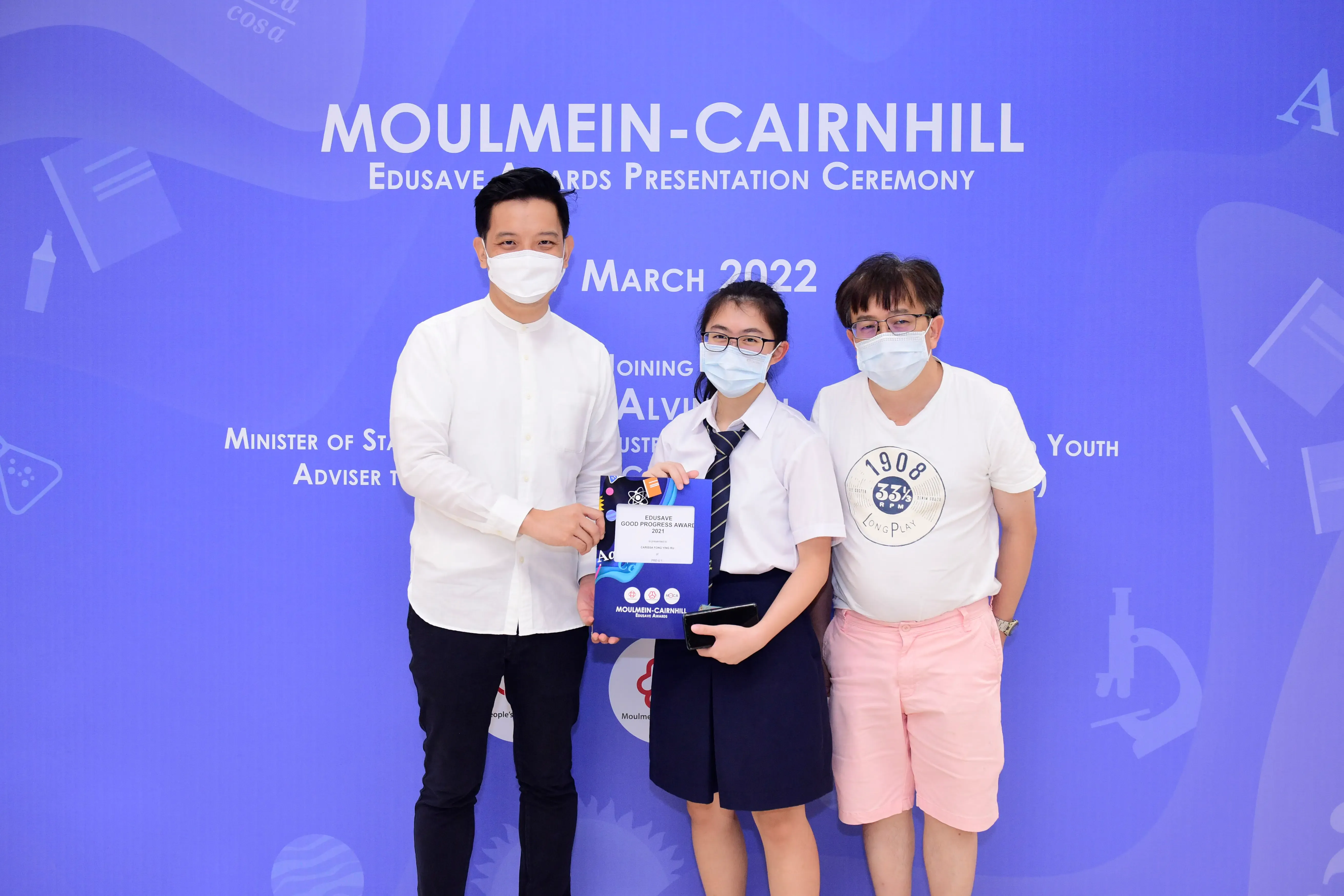 Three masked people pose at the Moulmein-Cairnhill Edusave Awards Ceremony in March 2022, with a student holding an award.