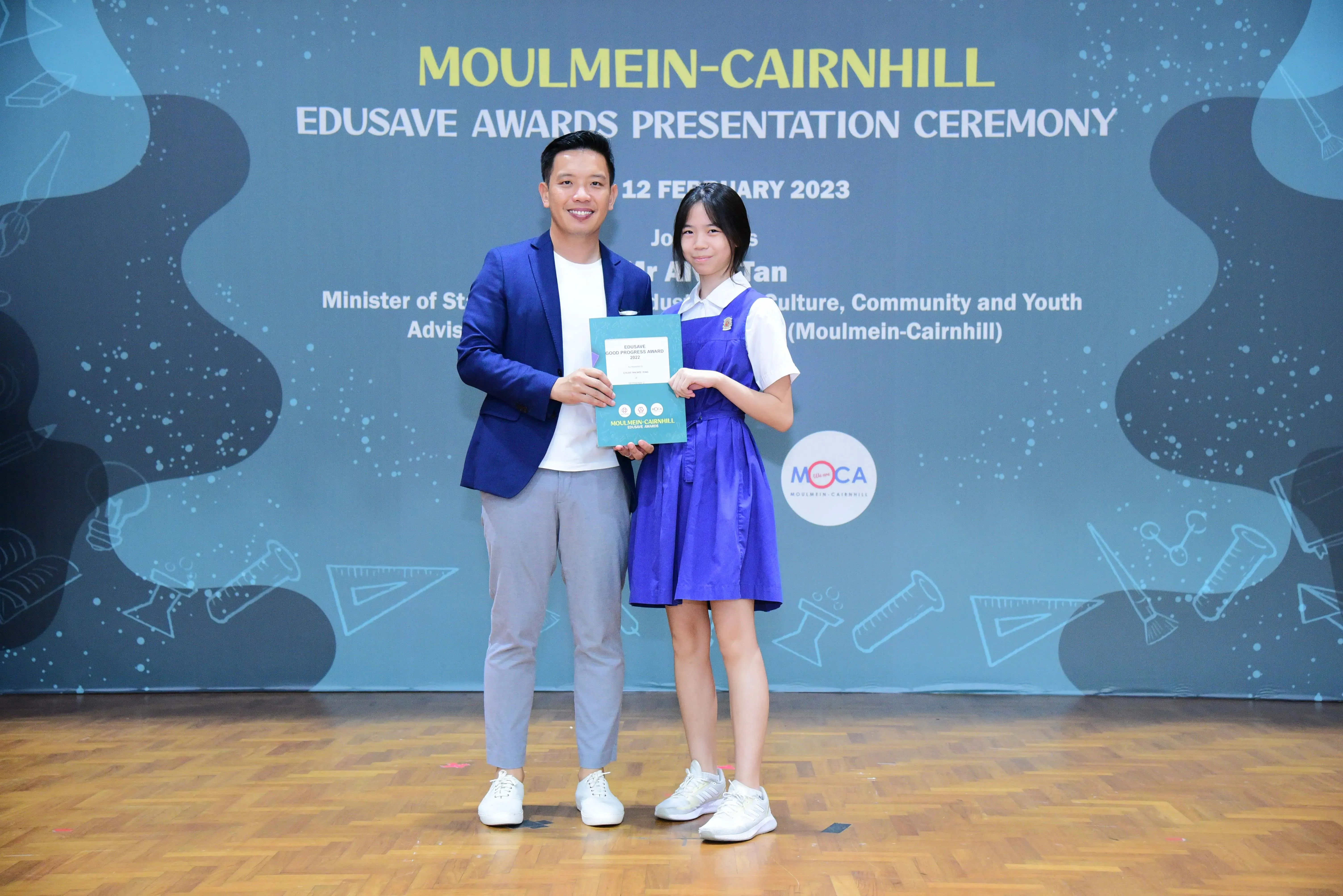 A man in a blue blazer and a girl in a blue school uniform hold an award at the Moulmein-Cairnhill Edusave ceremony.