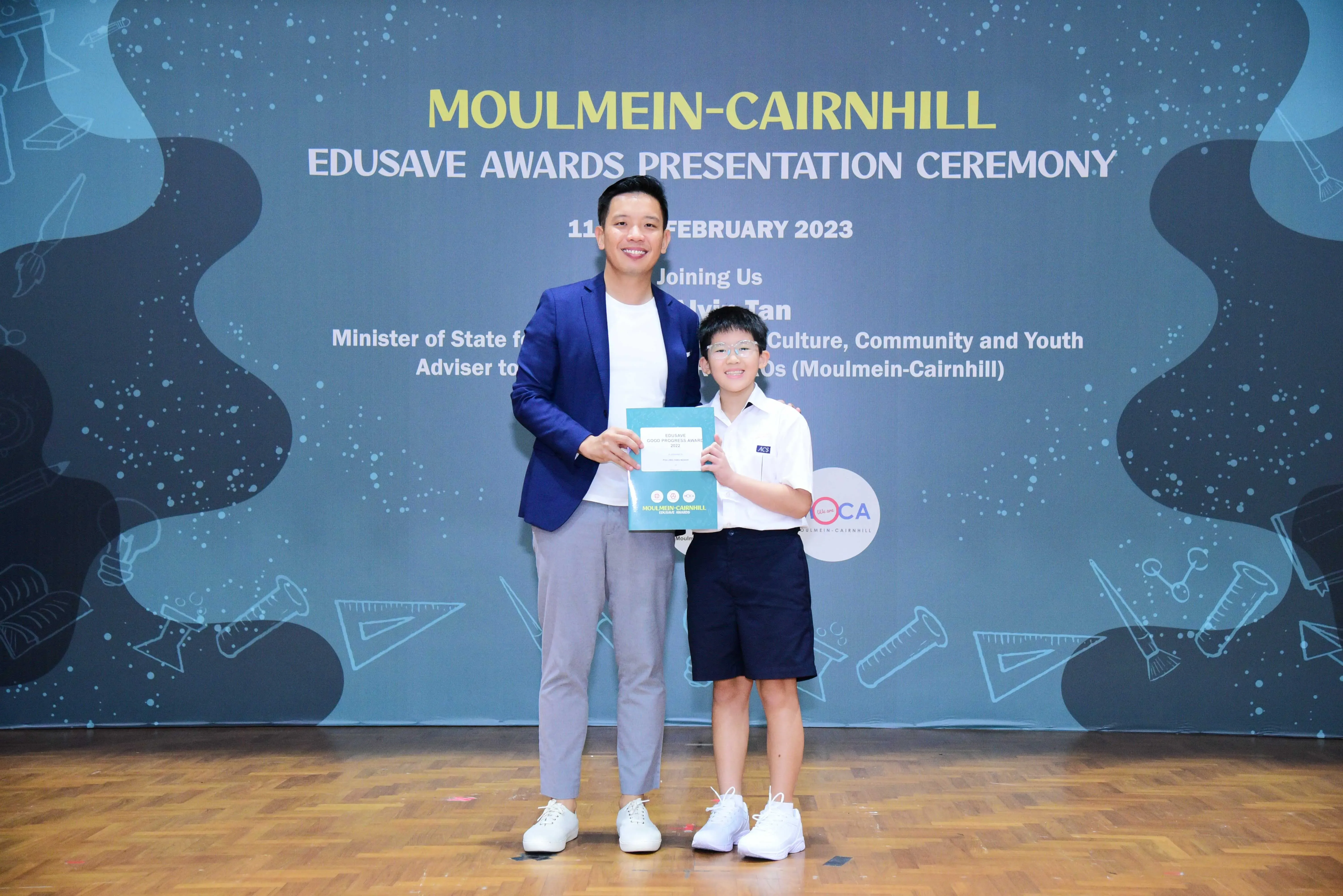 An adult in a blue blazer and a young boy in school uniform hold a certificate at the Moulmein-Cairnhill Edusave Awards 2023.