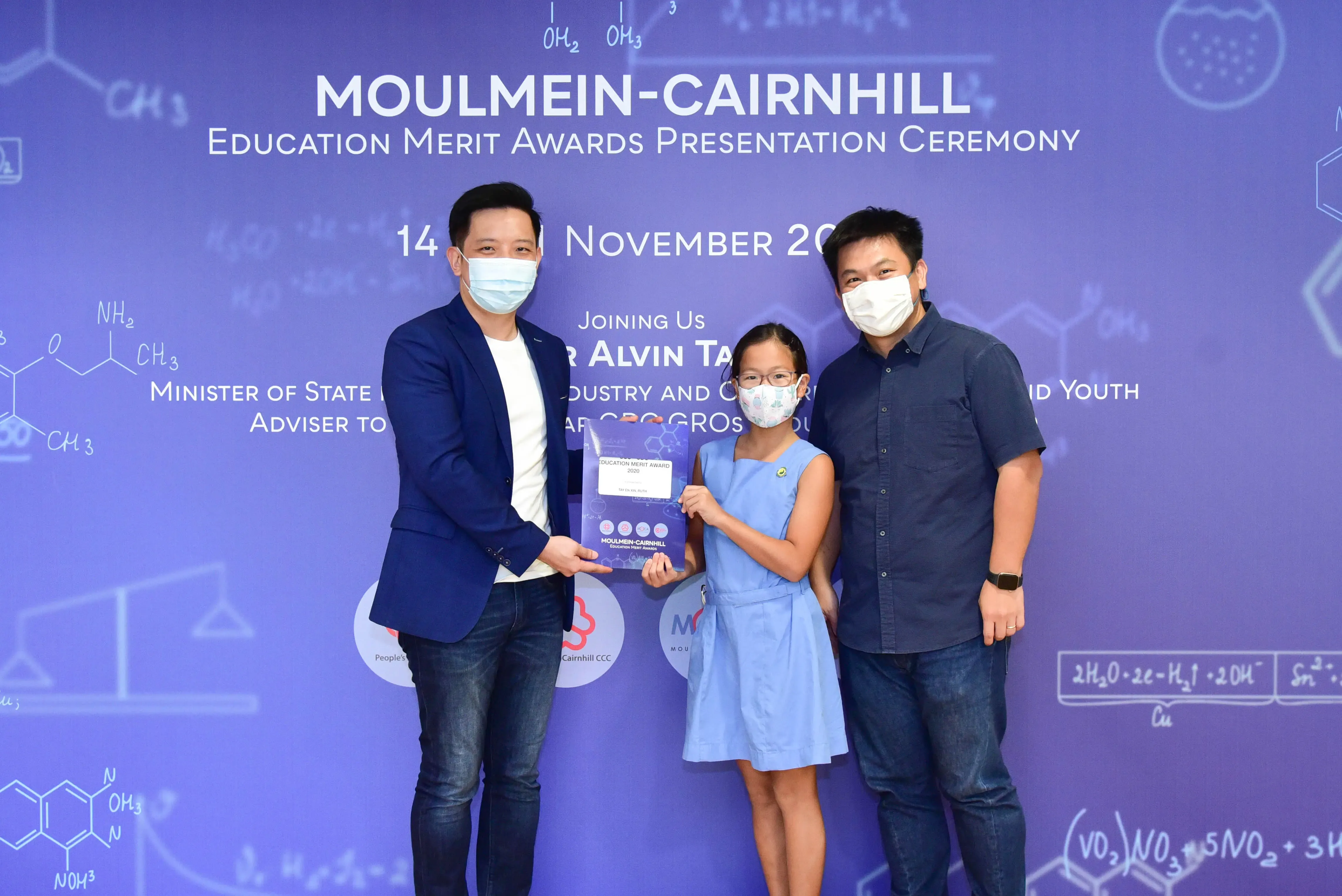 Two masked men and a masked girl hold an award plaque at the Moulmein-Cairnhill Education Merit Awards ceremony.