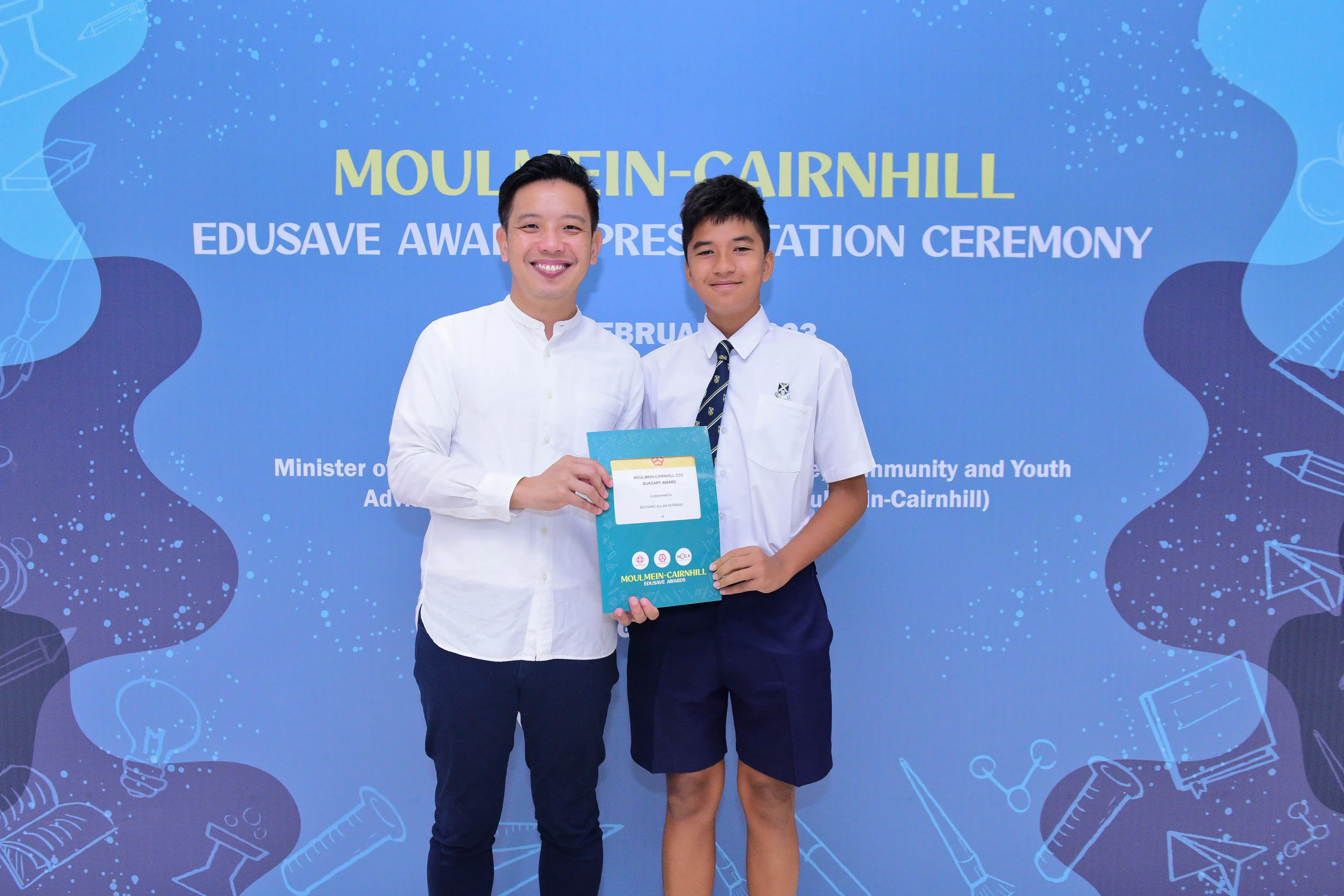 Two people hold a certificate at the Moulmein-Cairnhill Edusave Awards Presentation Ceremony backdrop.