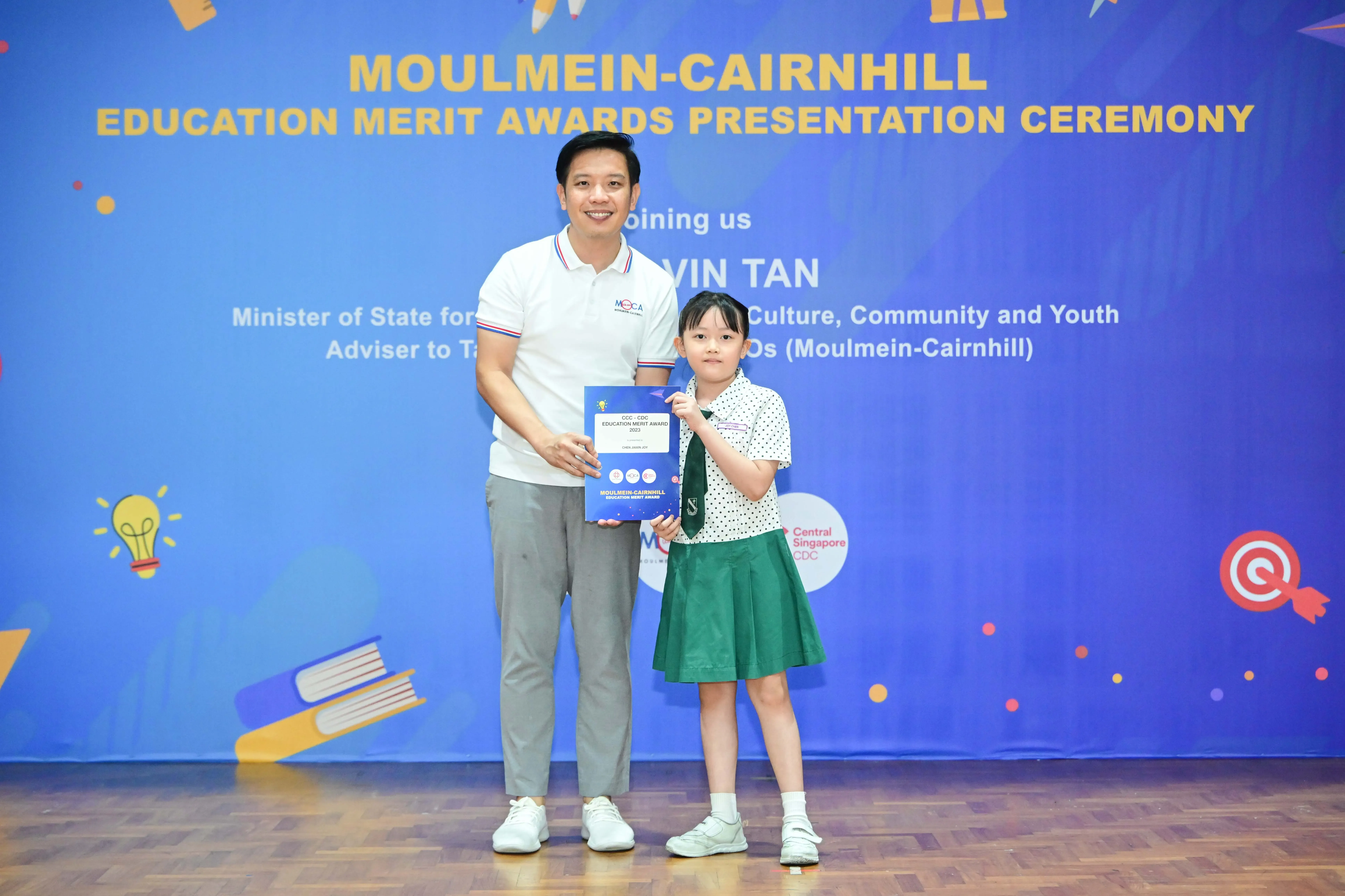 A man and a young girl in school uniform hold a merit award certificate at the Moulmein-Cairnhill Education Merit Awards ceremony.