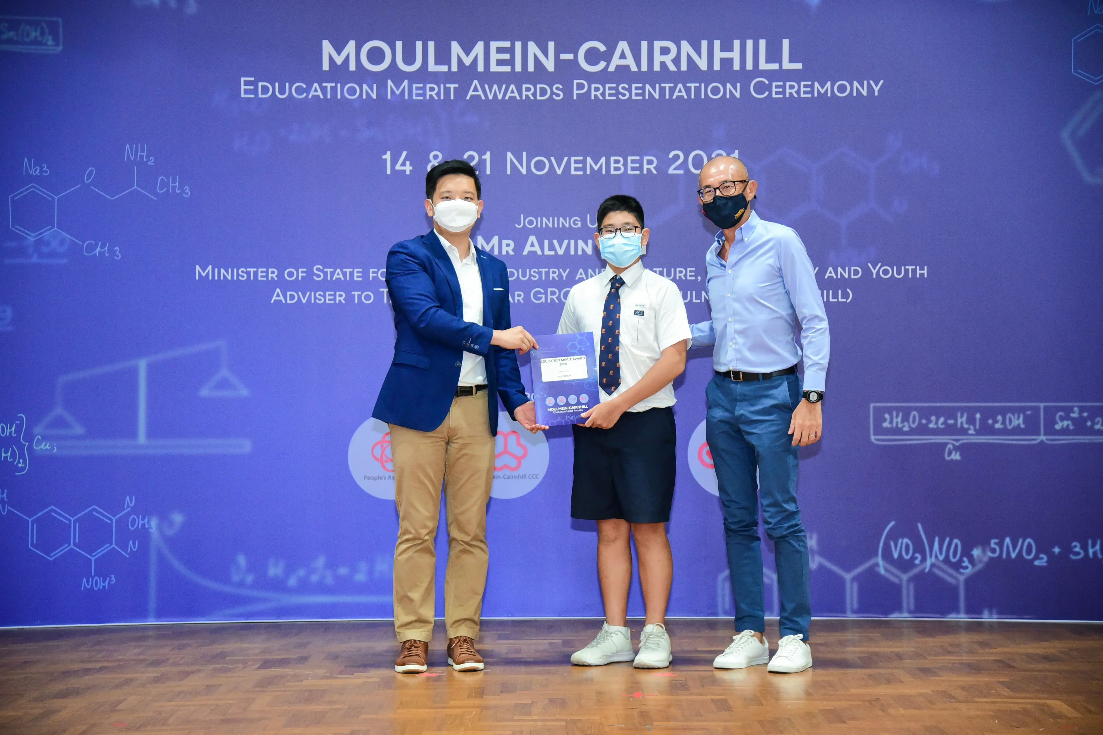 Two men and a student in uniform hold an award at the Moulmein-Cairnhill Education Merit Awards Ceremony, November 2021.