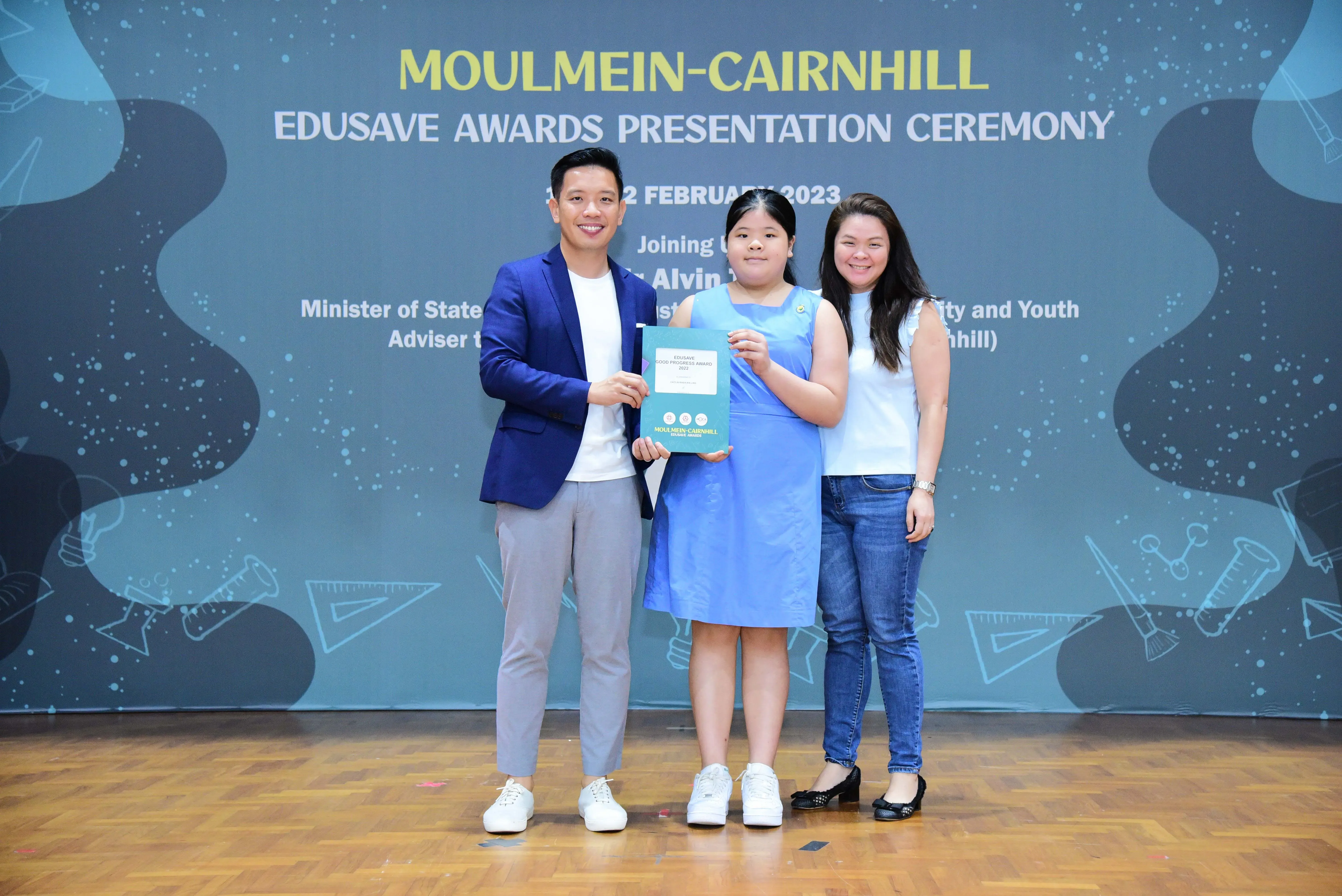 Three people smile on stage at the Moulmein-Cairnhill Edusave Awards Presentation Ceremony, with a girl holding a certificate.
