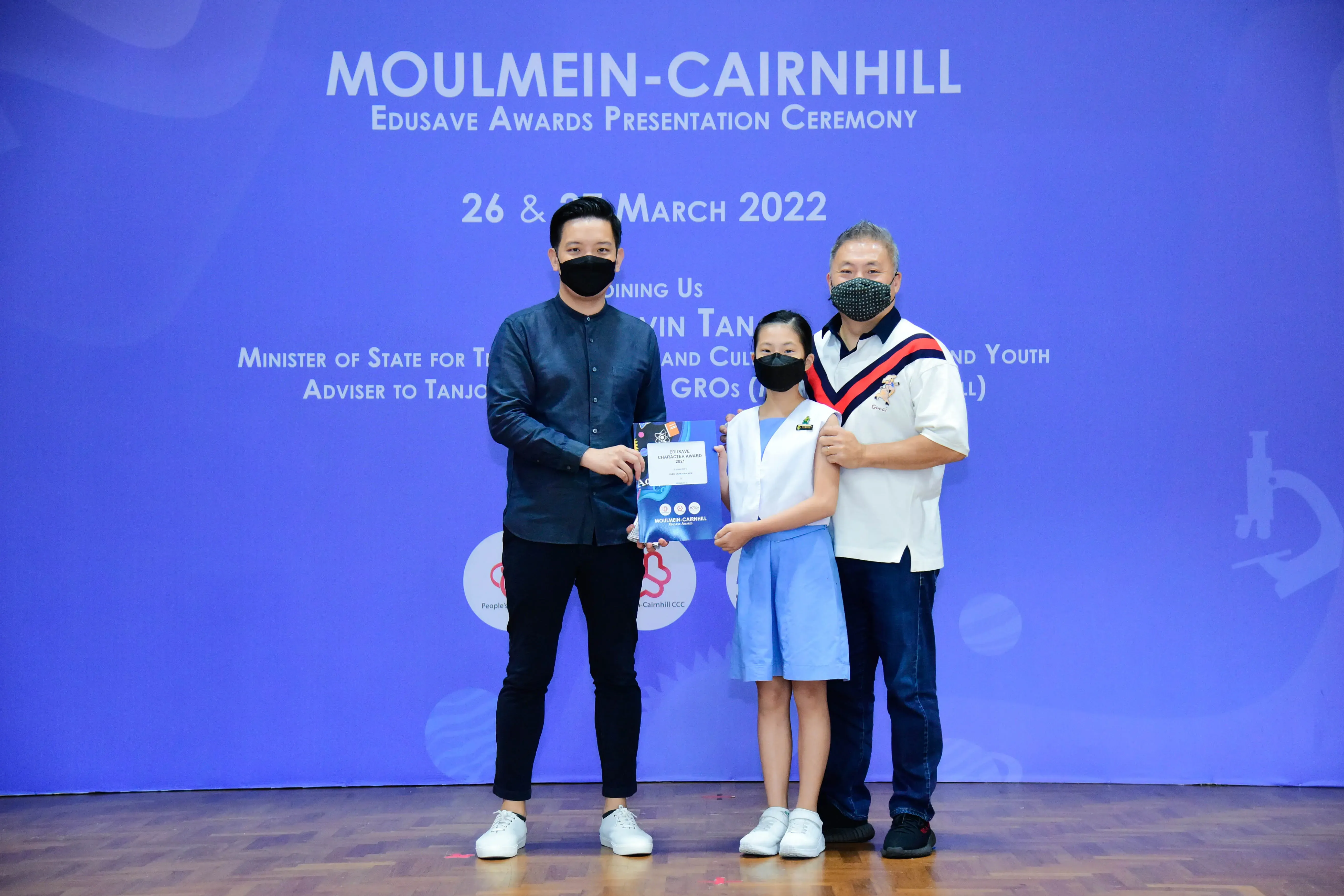 Two adults and a young girl in school uniform hold a certificate at the Moulmein-Cairnhill Edusave Awards Ceremony 2022.