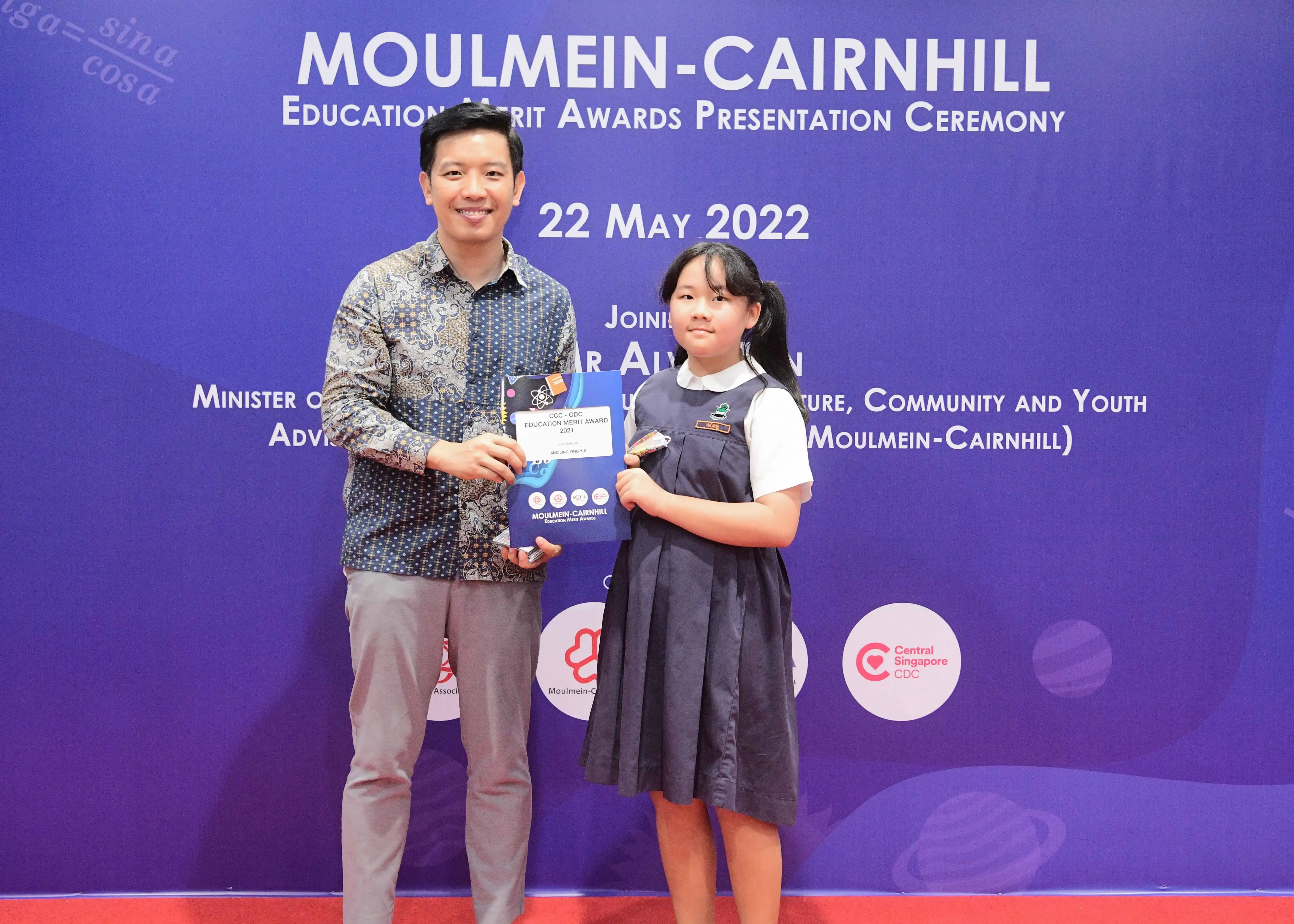 A man and a young girl in school uniform hold an award certificate at the Moulmein-Cairnhill Education Merit Awards ceremony.