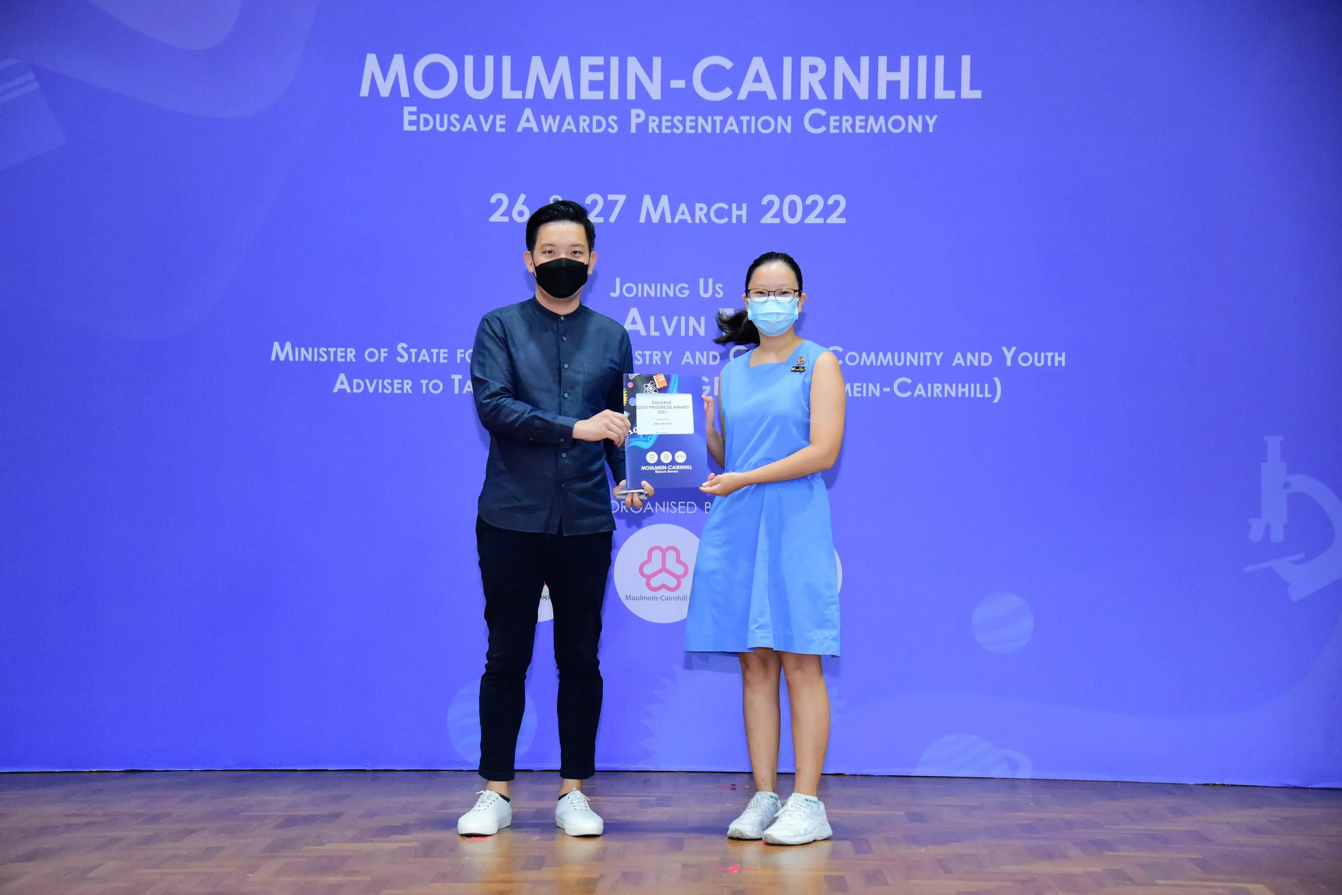 Two masked people hold a certificate on stage at the Moulmein-Cairnhill Edusave Awards Ceremony, March 2022.