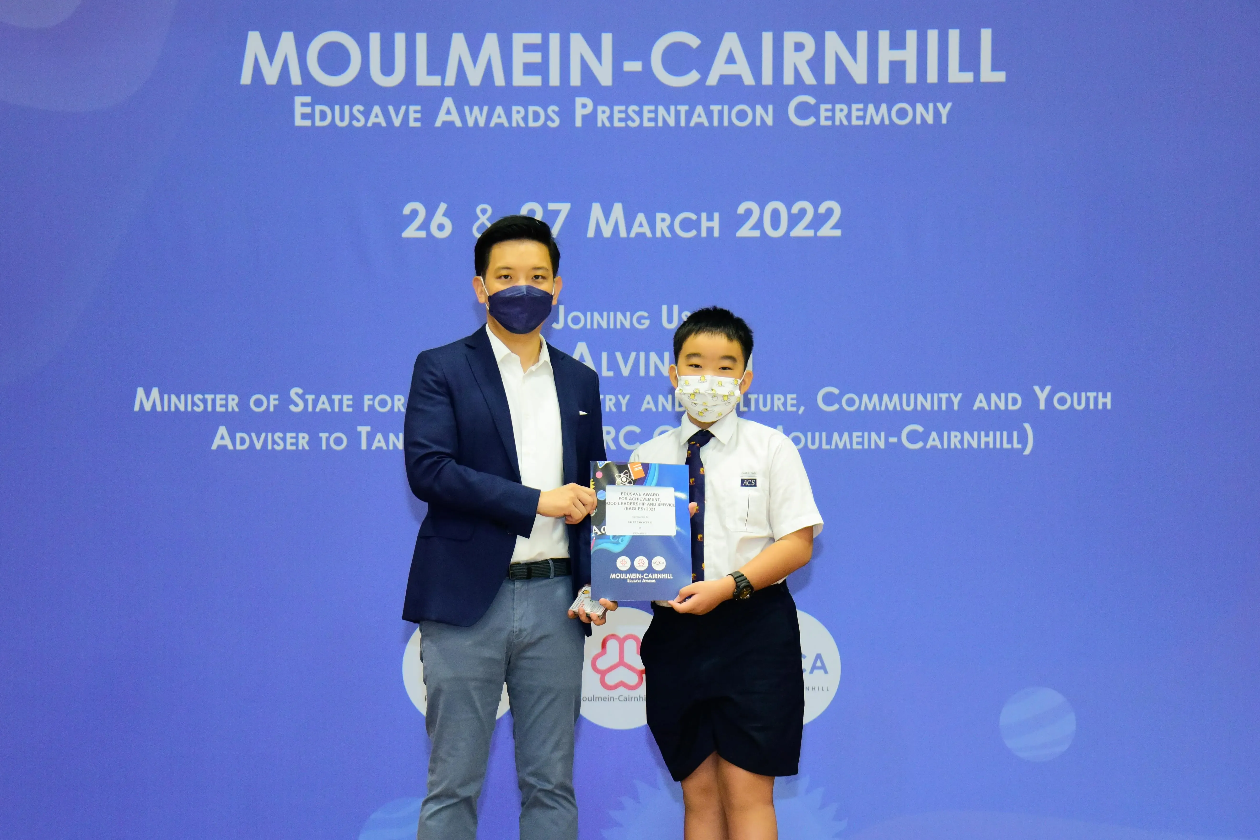 An adult and a young student in uniform hold an award together at the Moulmein-Cairnhill Edusave Awards Ceremony, March 2022.
