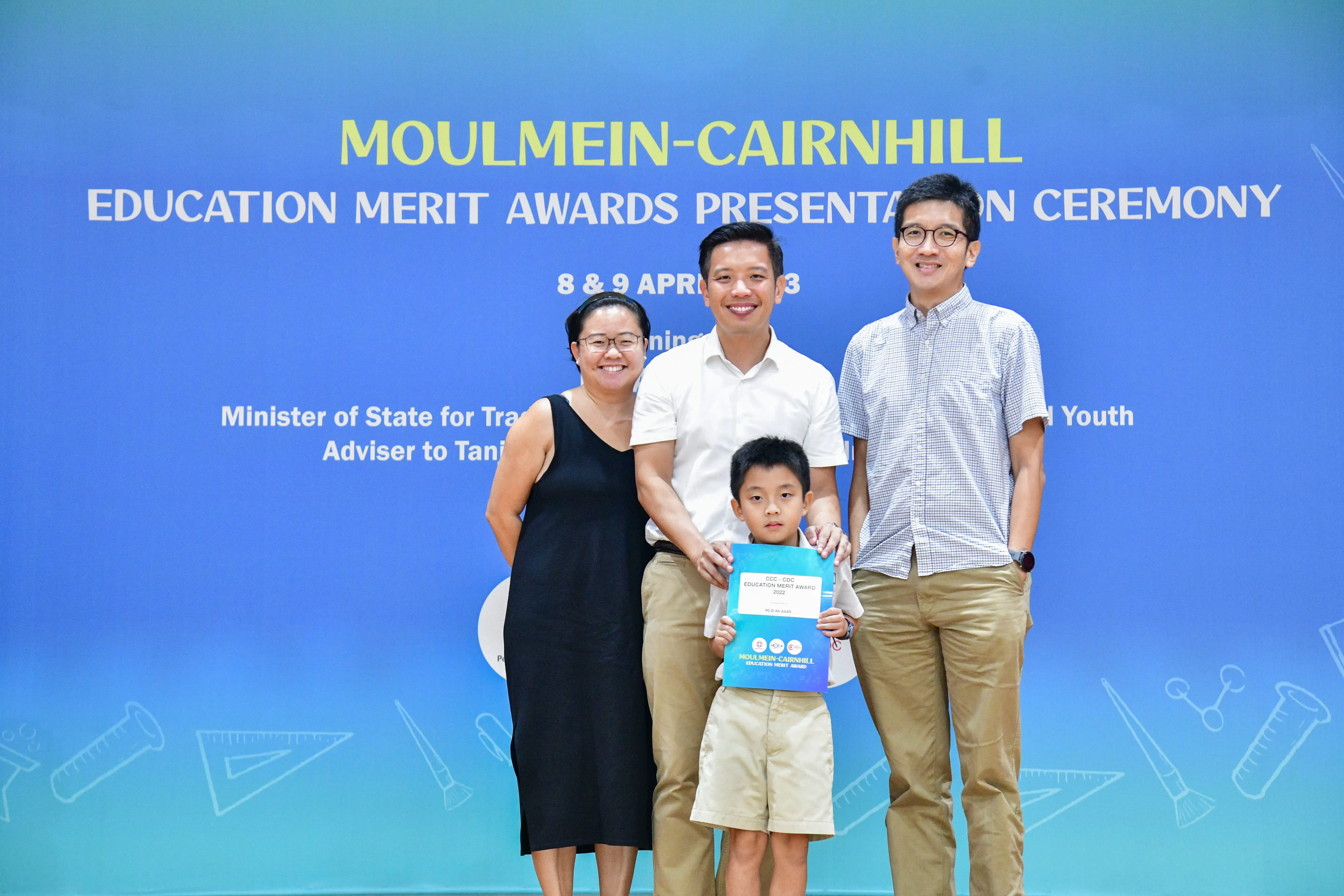 Three adults and a young boy holding a certificate pose before a Moulmein-Cairnhill Education Merit Awards banner.