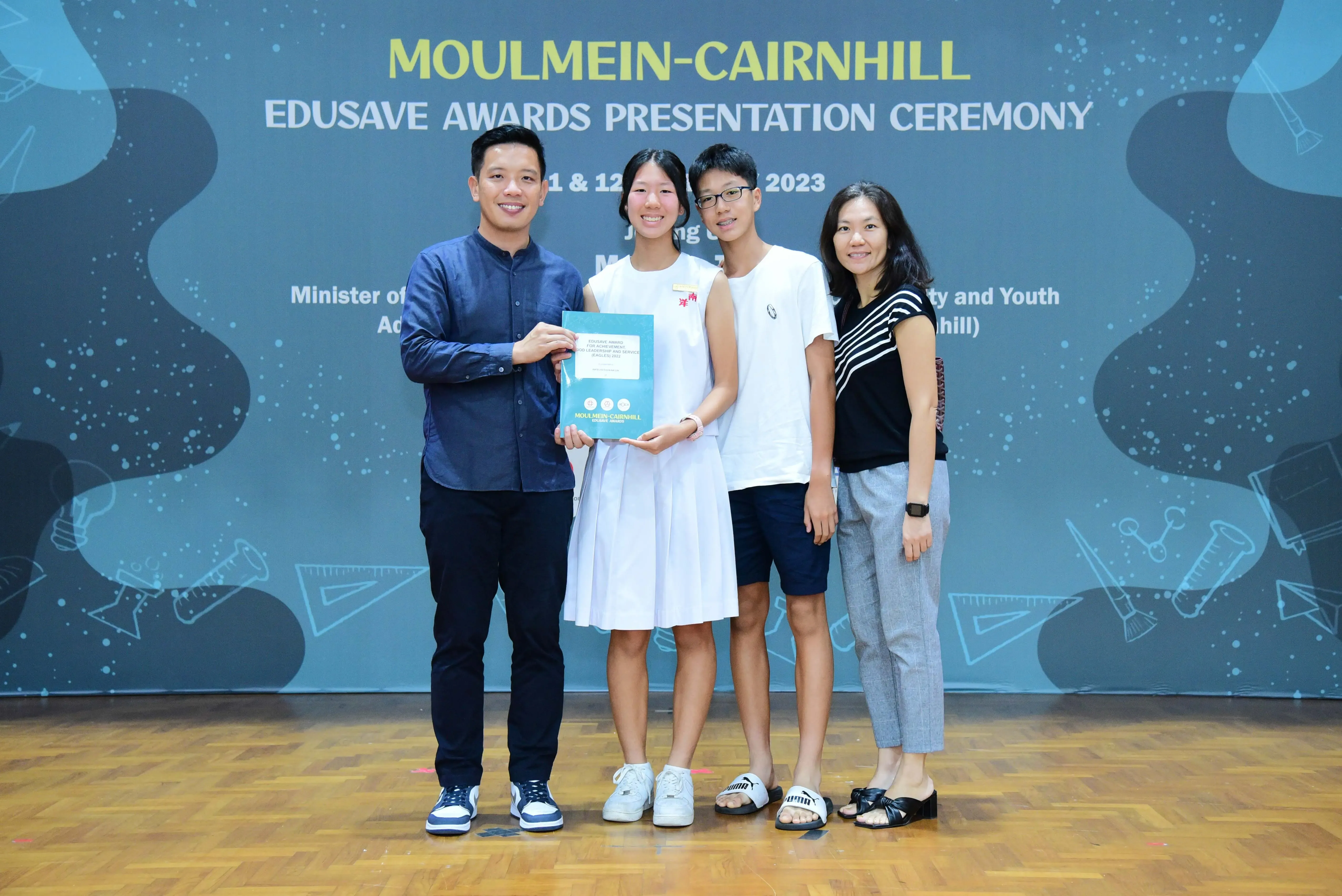 Four people pose holding a booklet at the Moulmein-Cairnhill Edusave Awards Presentation Ceremony 2023.
