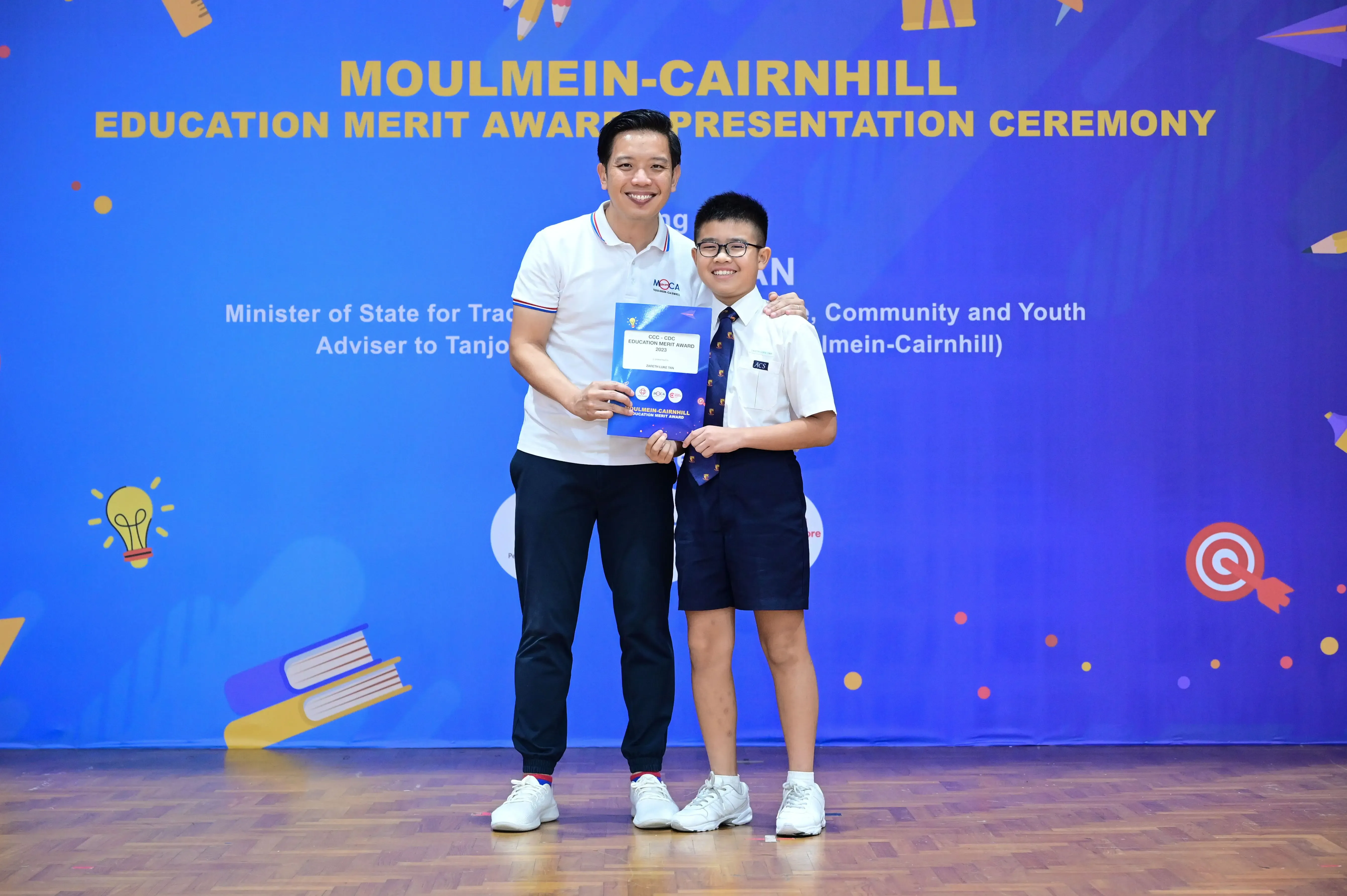 An adult and a young boy in school uniform hold an award certificate at the Moulmein-Cairnhill Education Merit Award Ceremony.