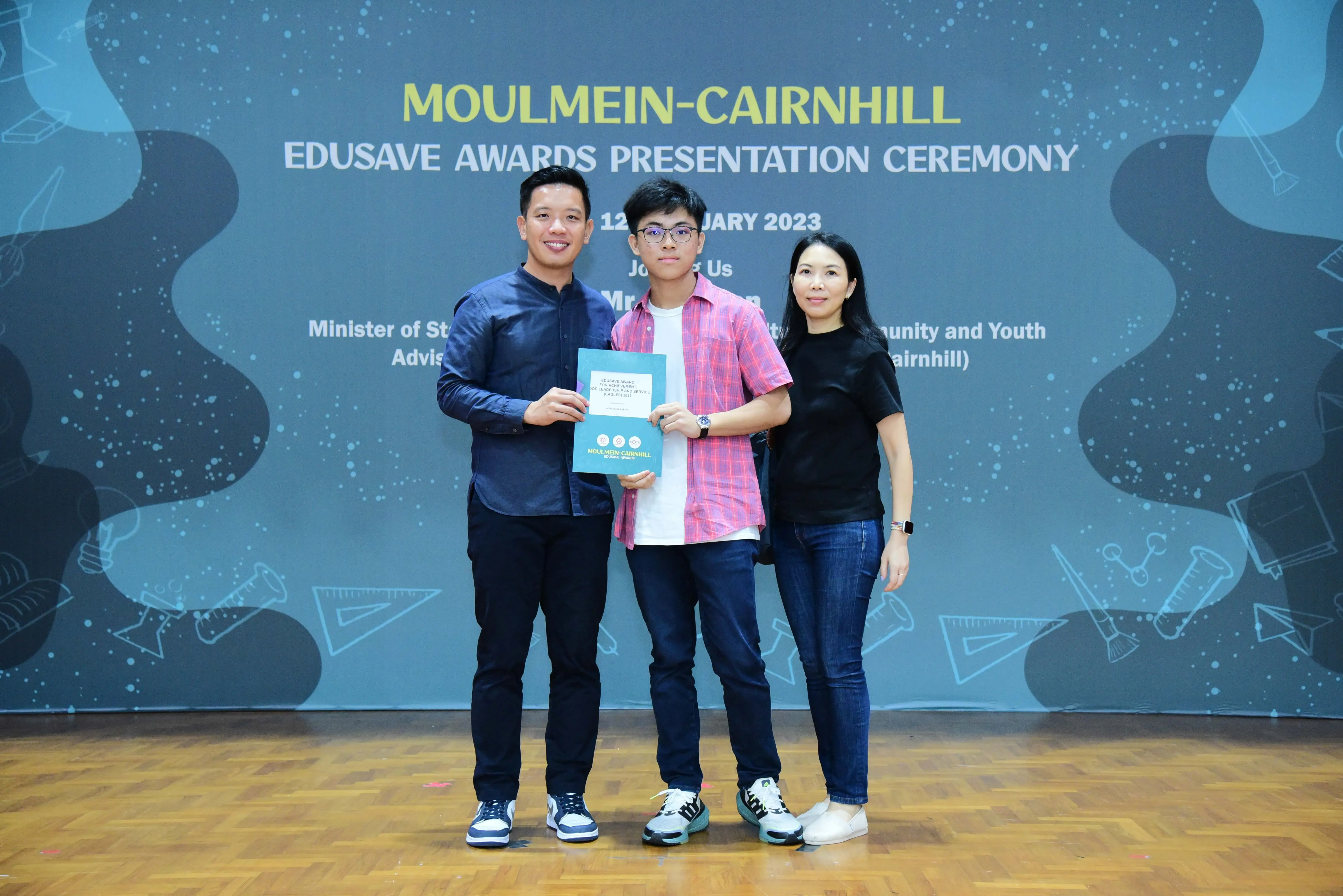Three people stand together on a stage at the Moulmein-Cairnhill Edusave Awards Presentation Ceremony 2023.