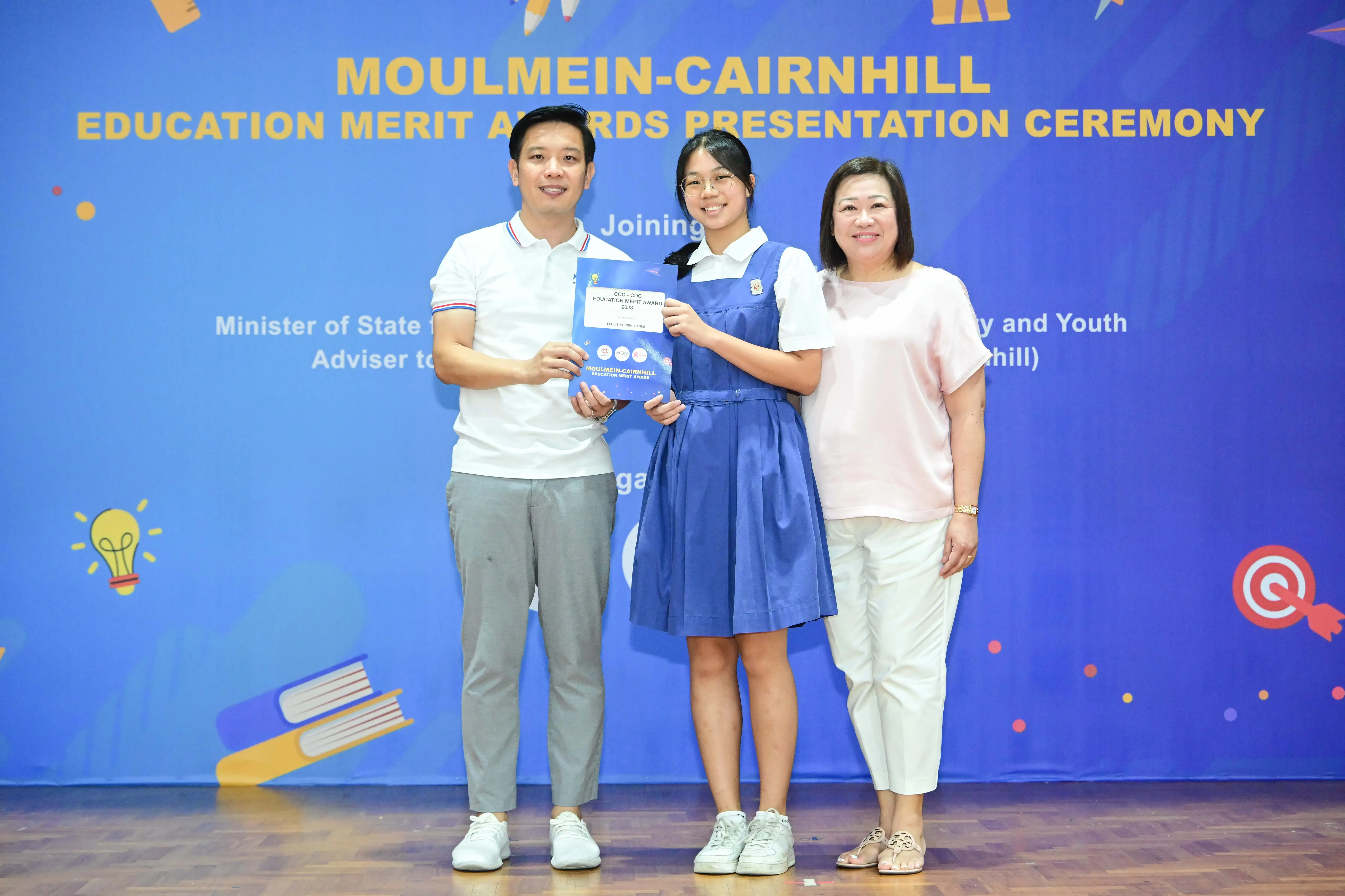 Three people pose smiling at the Moulmein-Cairnhill Education Merit Awards Presentation Ceremony, student holding a certificate.