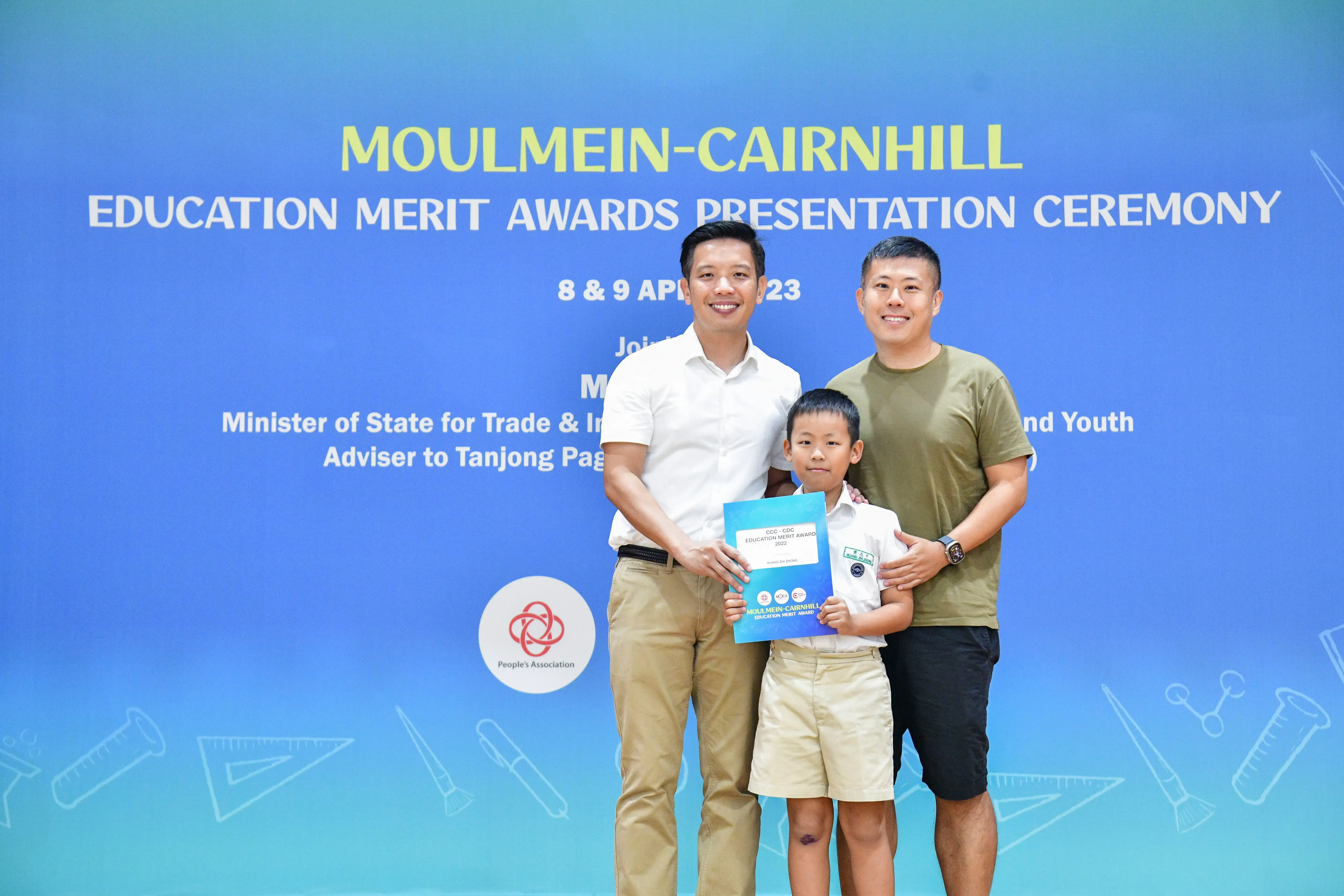 Two men and a young boy hold an award certificate in front of a Moulmein-Cairnhill Education Merit Awards banner.
