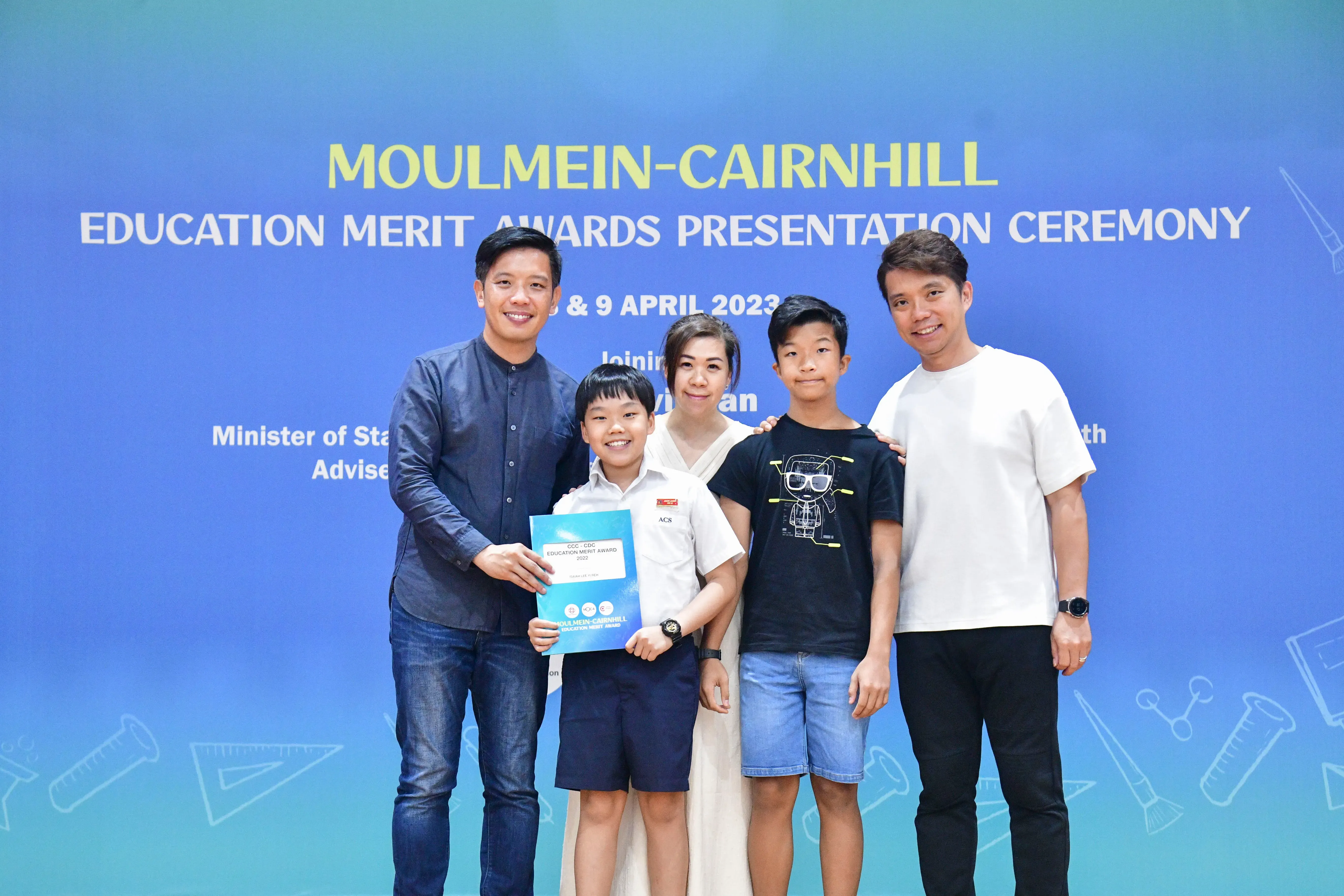 Five people pose before a Moulmein-Cairnhill Education Merit Awards Presentation Ceremony banner, with a boy holding a certificate.