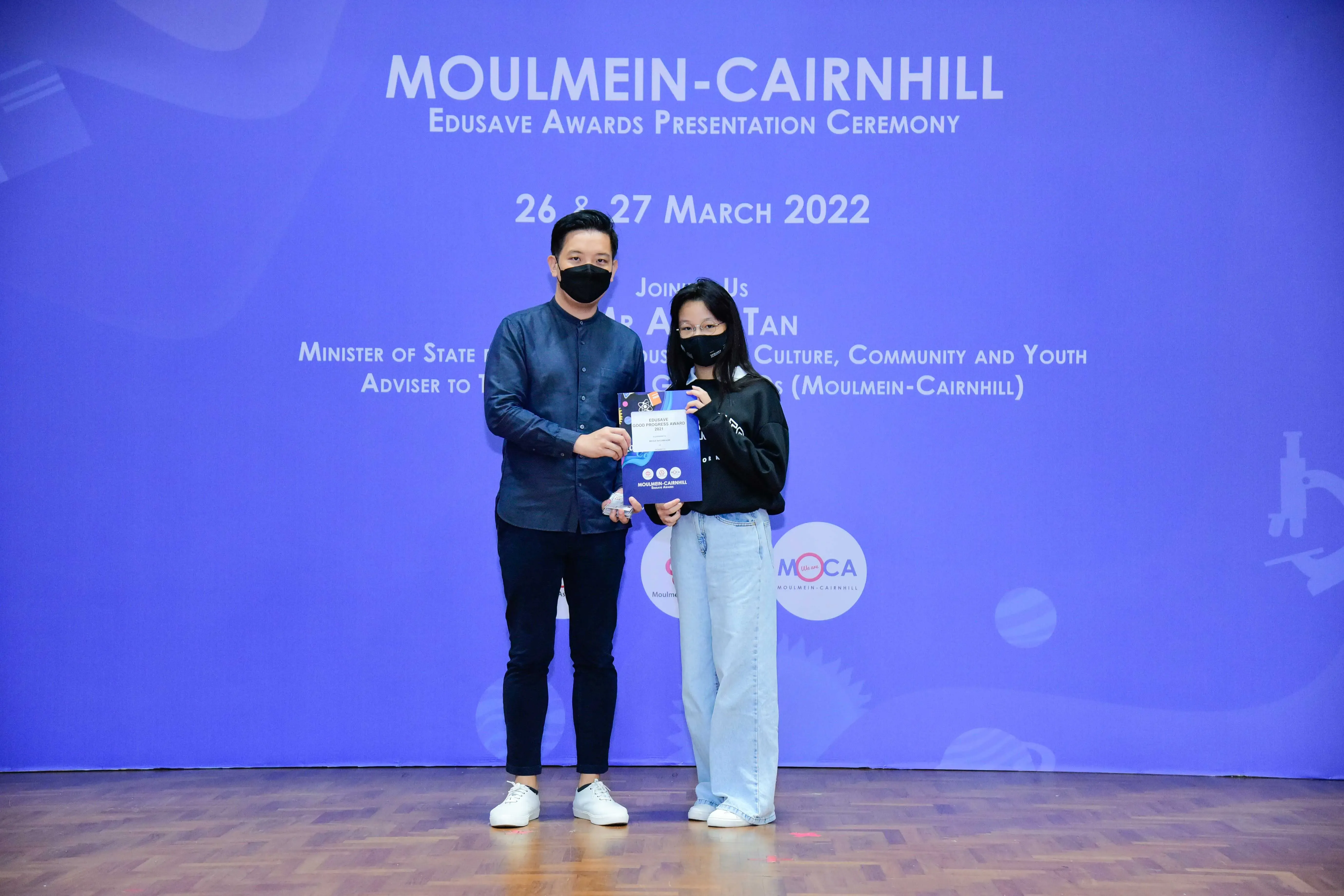 Two masked people hold an award certificate at the Moulmein-Cairnhill Edusave Awards Ceremony, March 2022.
