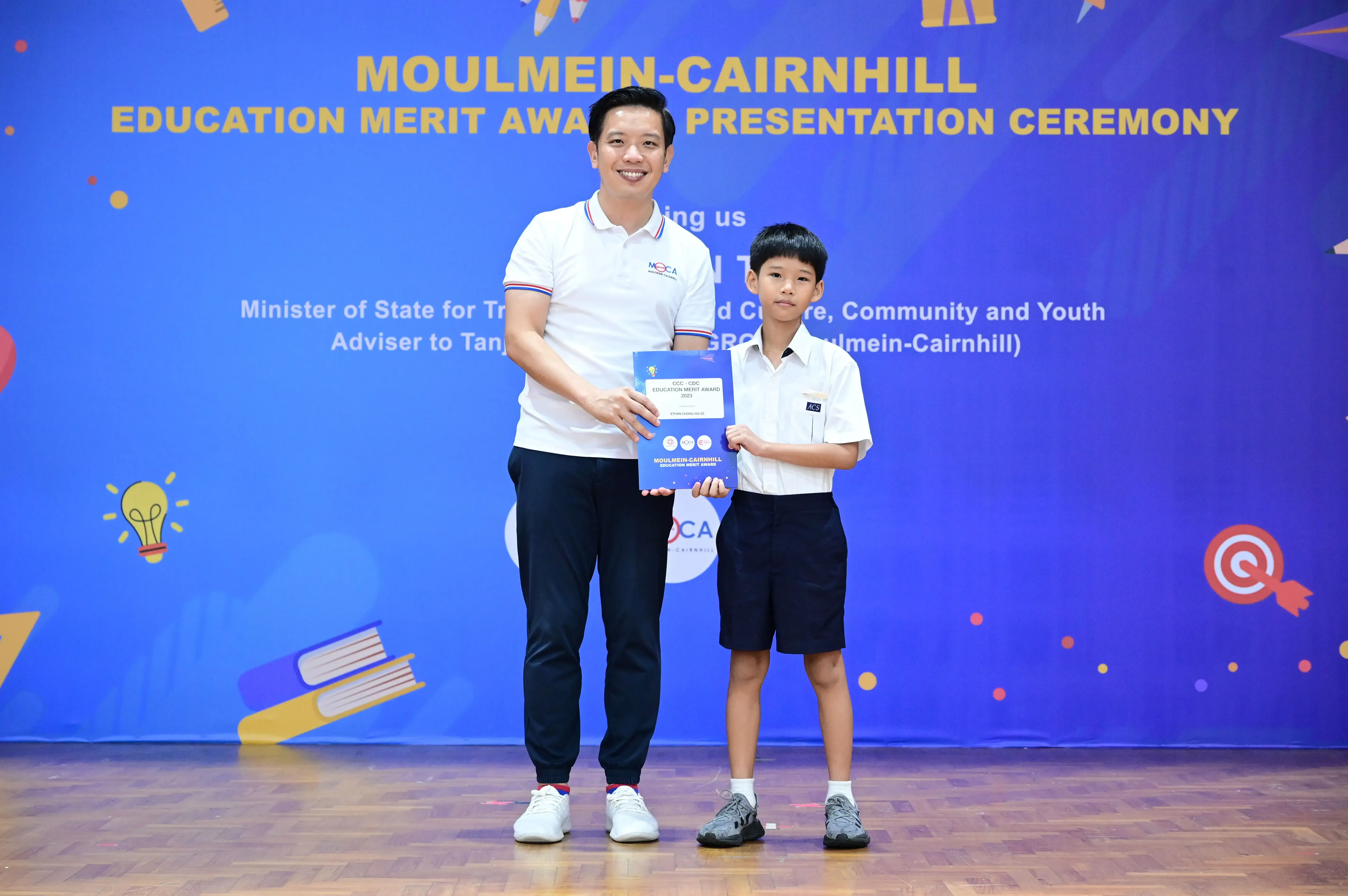Adult man and young boy hold an award certificate on stage at Moulmein-Cairnhill Education Merit Award Ceremony.