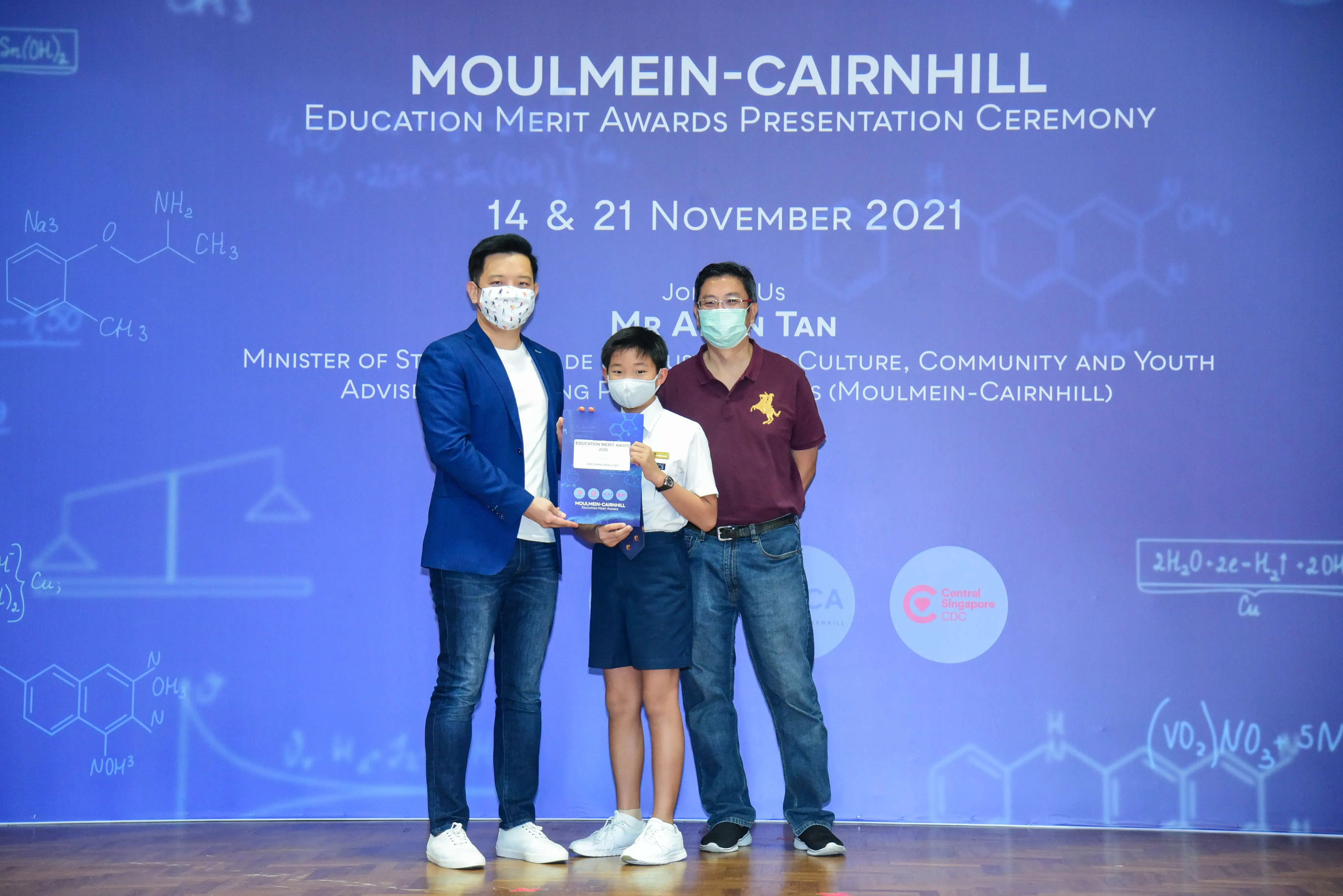Two masked adults and a young boy holding an award at the Moulmein-Cairnhill Education Merit Awards Ceremony, November 2021.