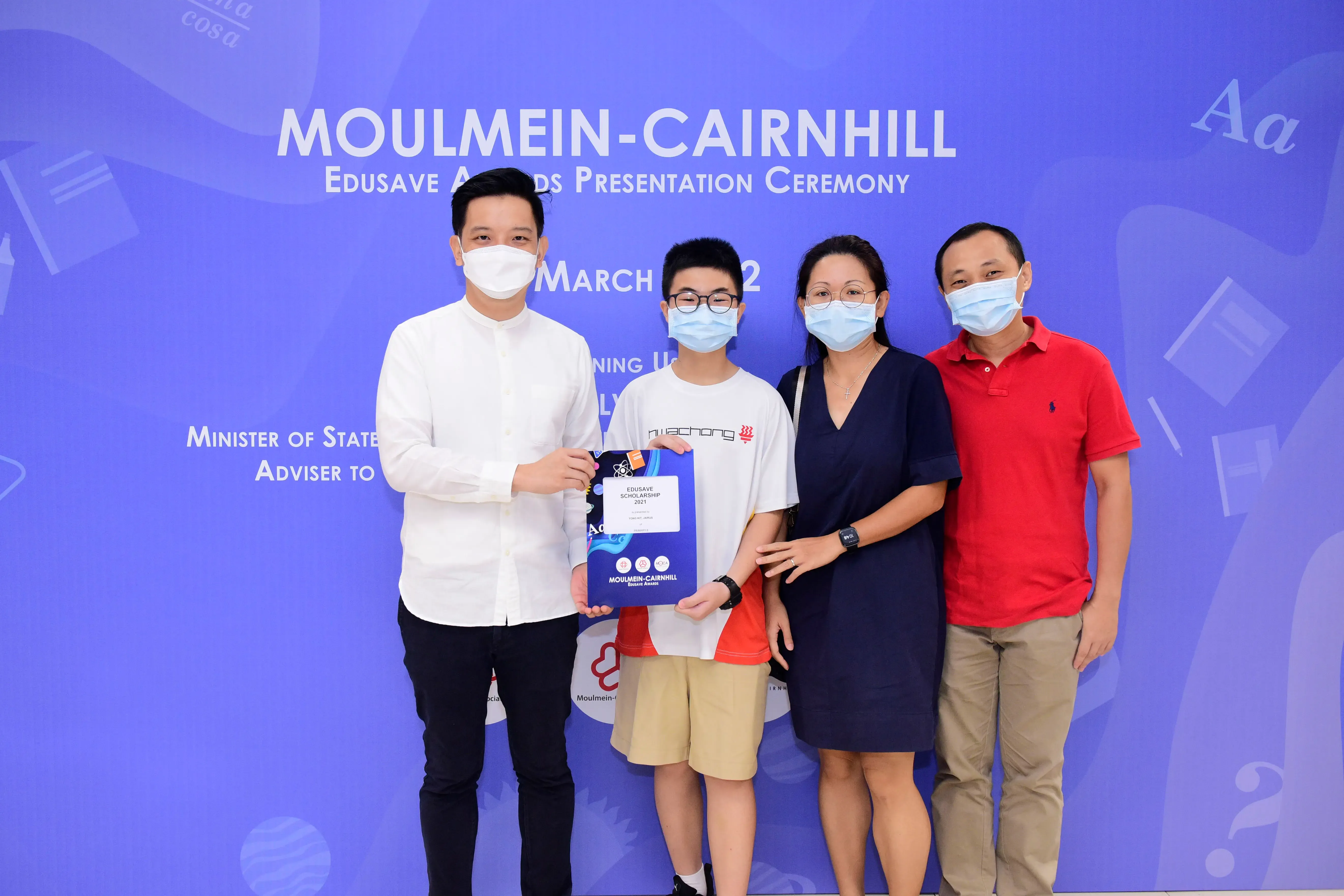 Four masked people pose at the Moulmein-Cairnhill Edusave Awards Presentation Ceremony, with a boy holding a scholarship folder.
