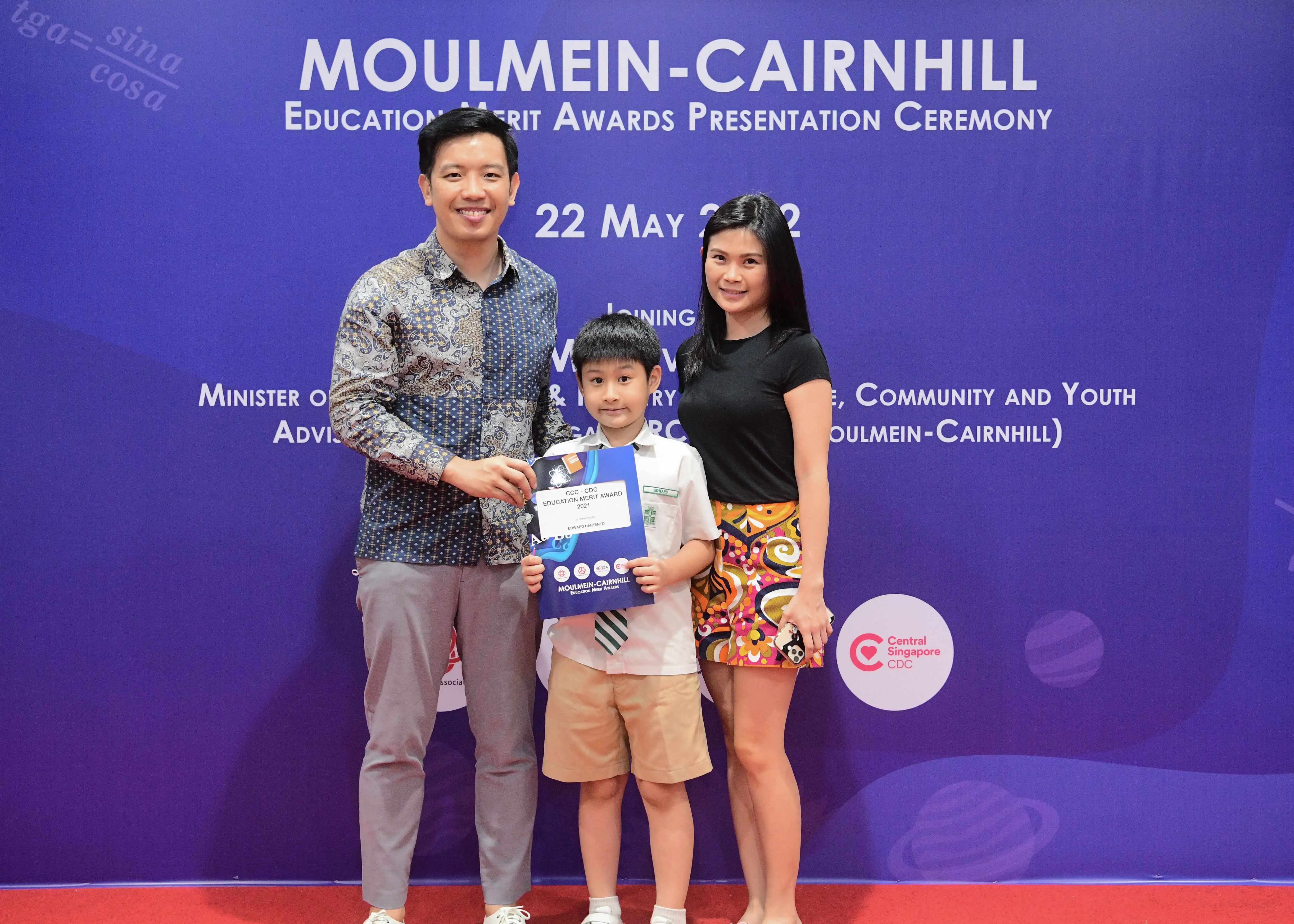 A man and woman pose with a young boy holding an award certificate at the Moulmein-Cairnhill Education Merit Awards ceremony.