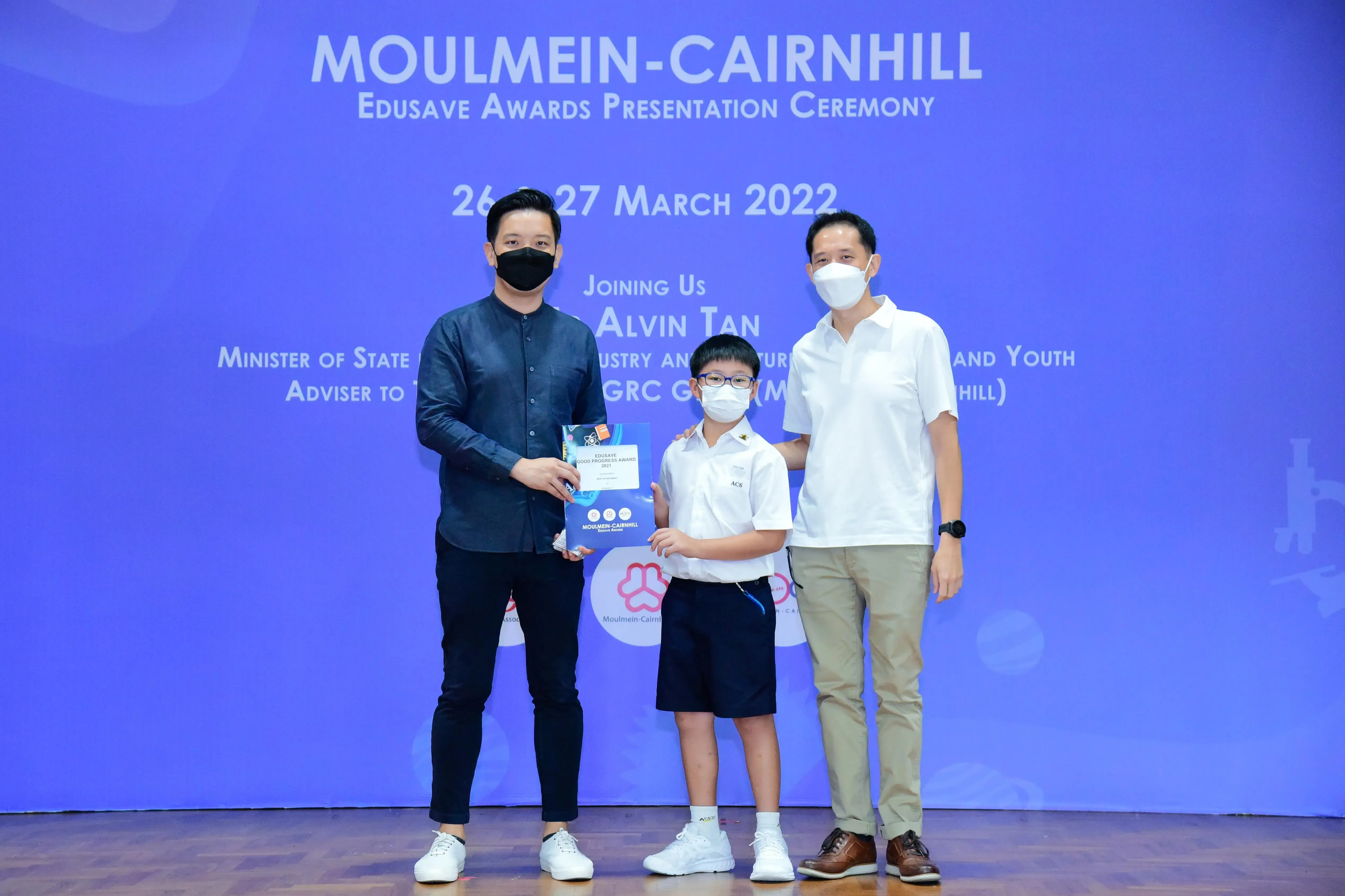Two masked adults and a masked child holding a certificate pose at the Moulmein-Cairnhill Edusave Awards Ceremony 2022.