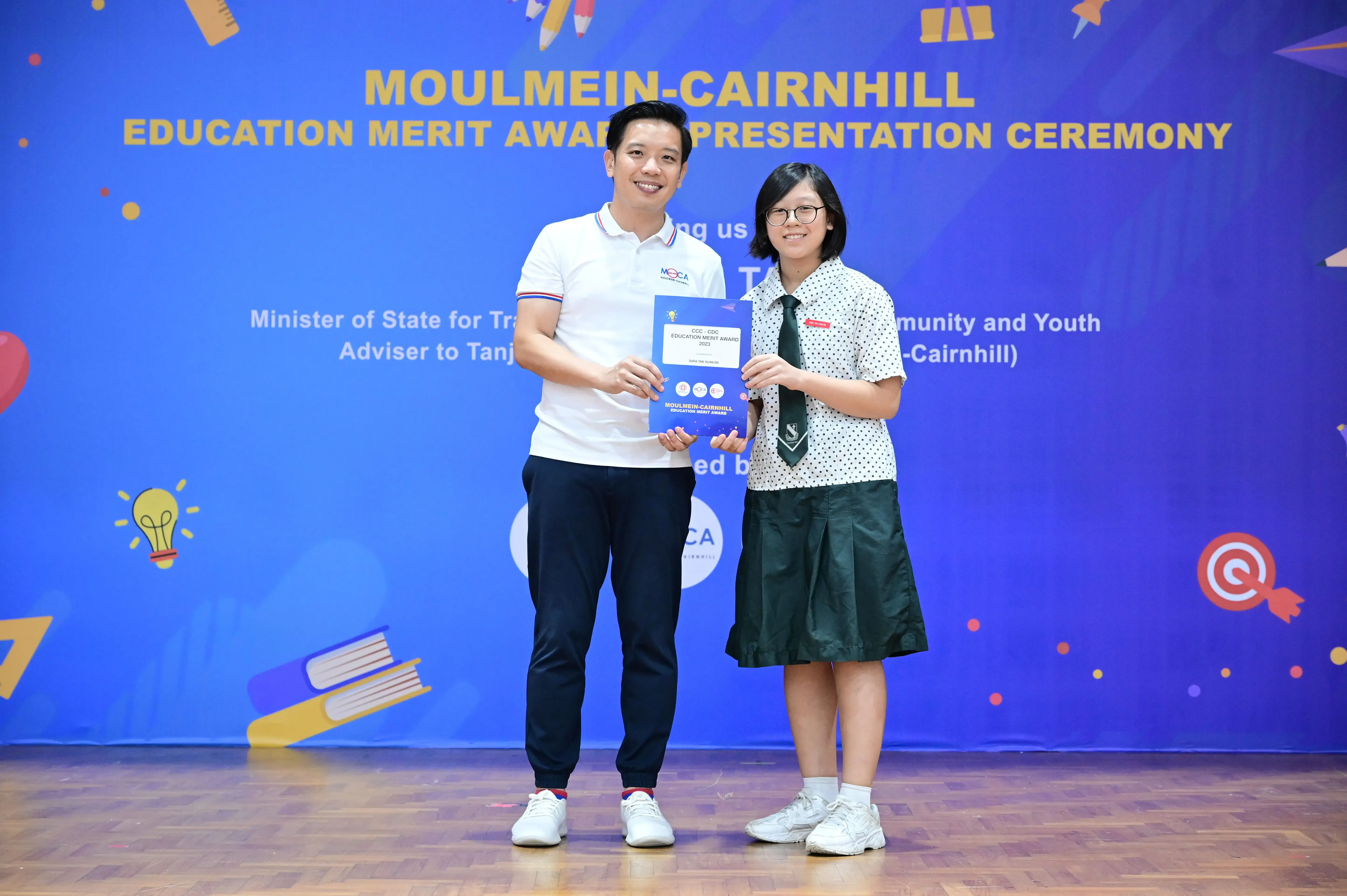 A man and a uniformed student hold a certificate at the Moulmein-Cairnhill Education Merit Award Presentation Ceremony.