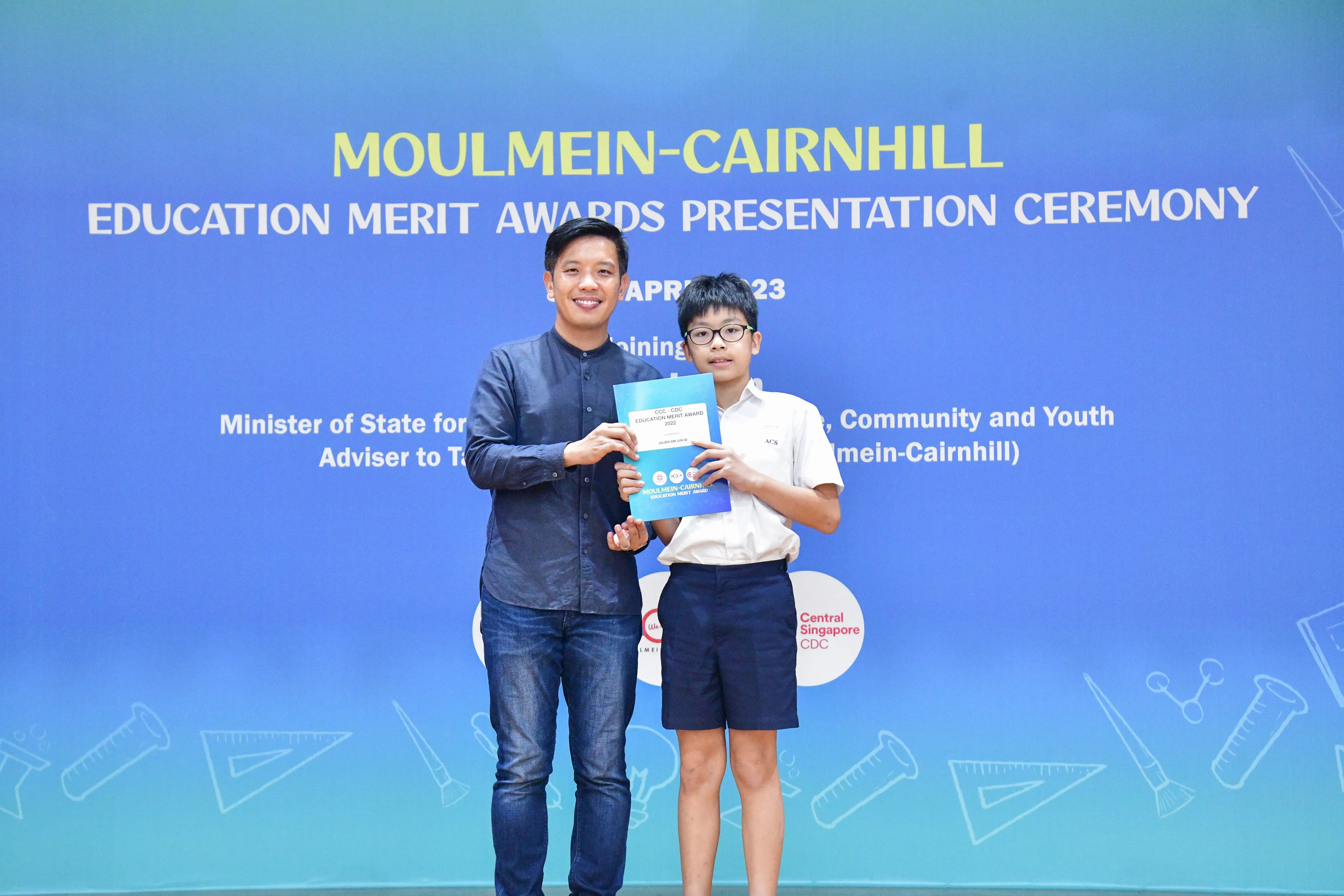 An adult and a young boy hold a certificate together at the Moulmein-Cairnhill Education Merit Awards Presentation Ceremony.