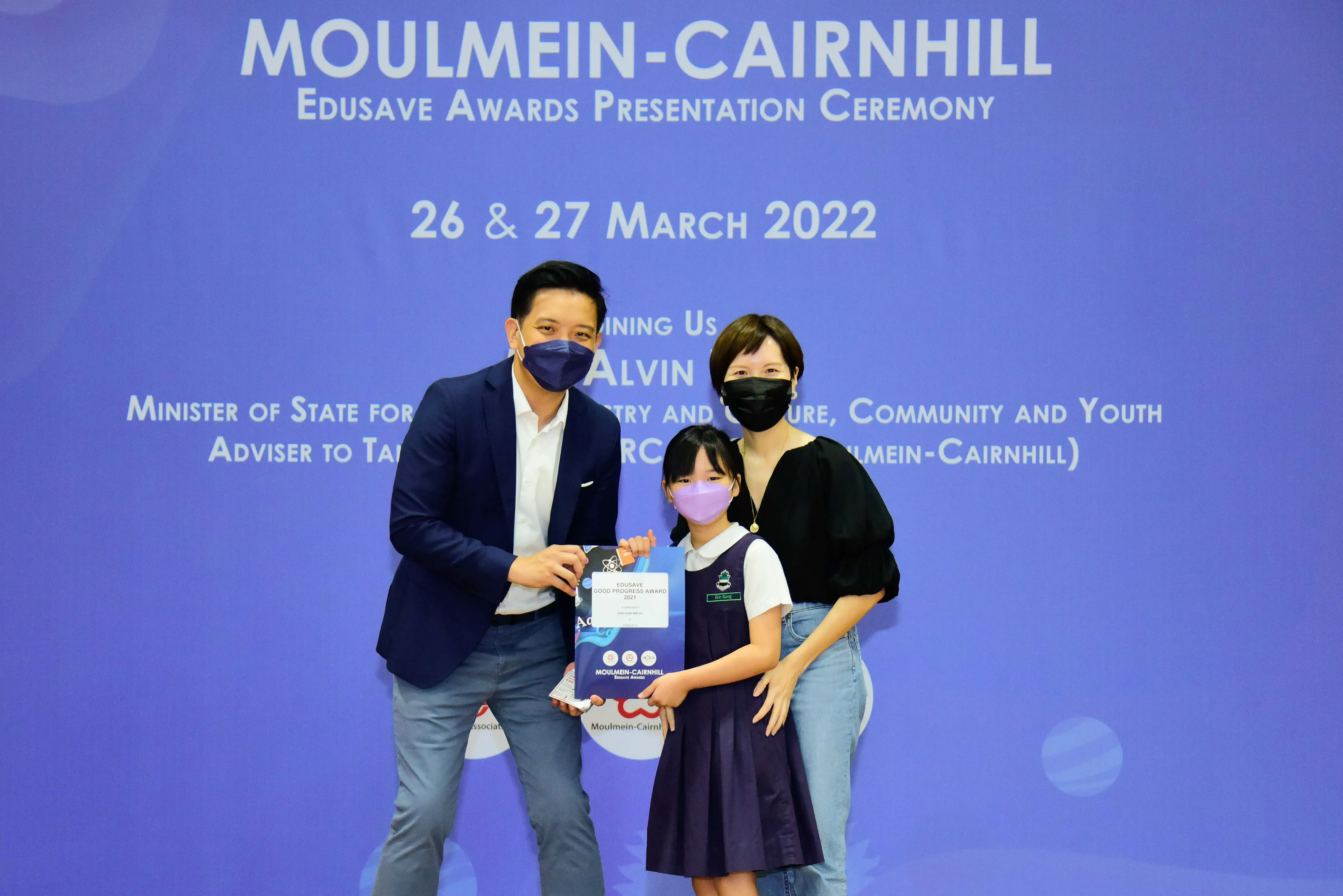 Two adults and a young girl in school uniform hold an award at the Moulmein-Cairnhill Edusave Awards Ceremony 2022.