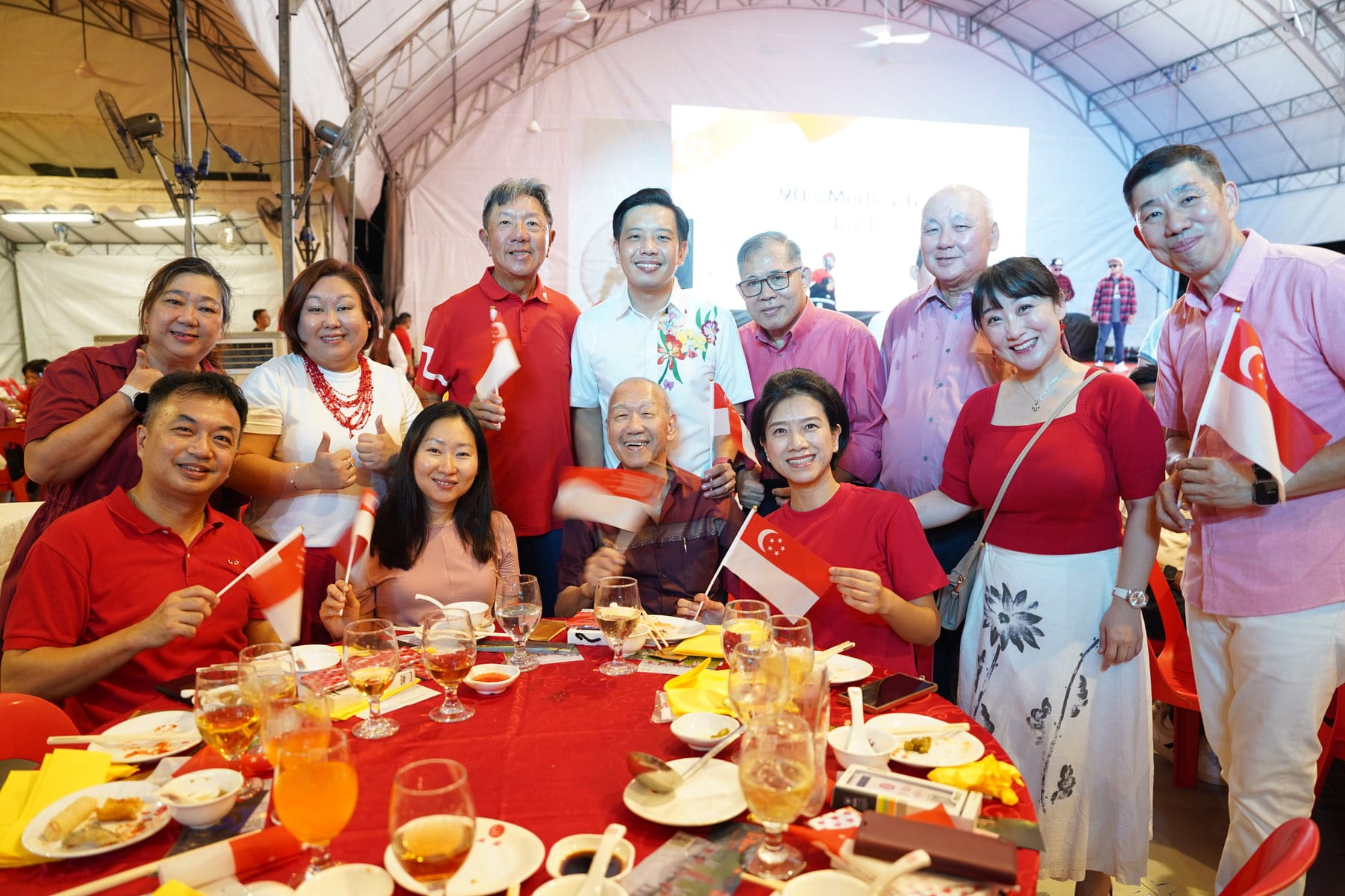 Group of smiling people in red and white clothing holding Singapore flags at a festive dinner under a tent.
