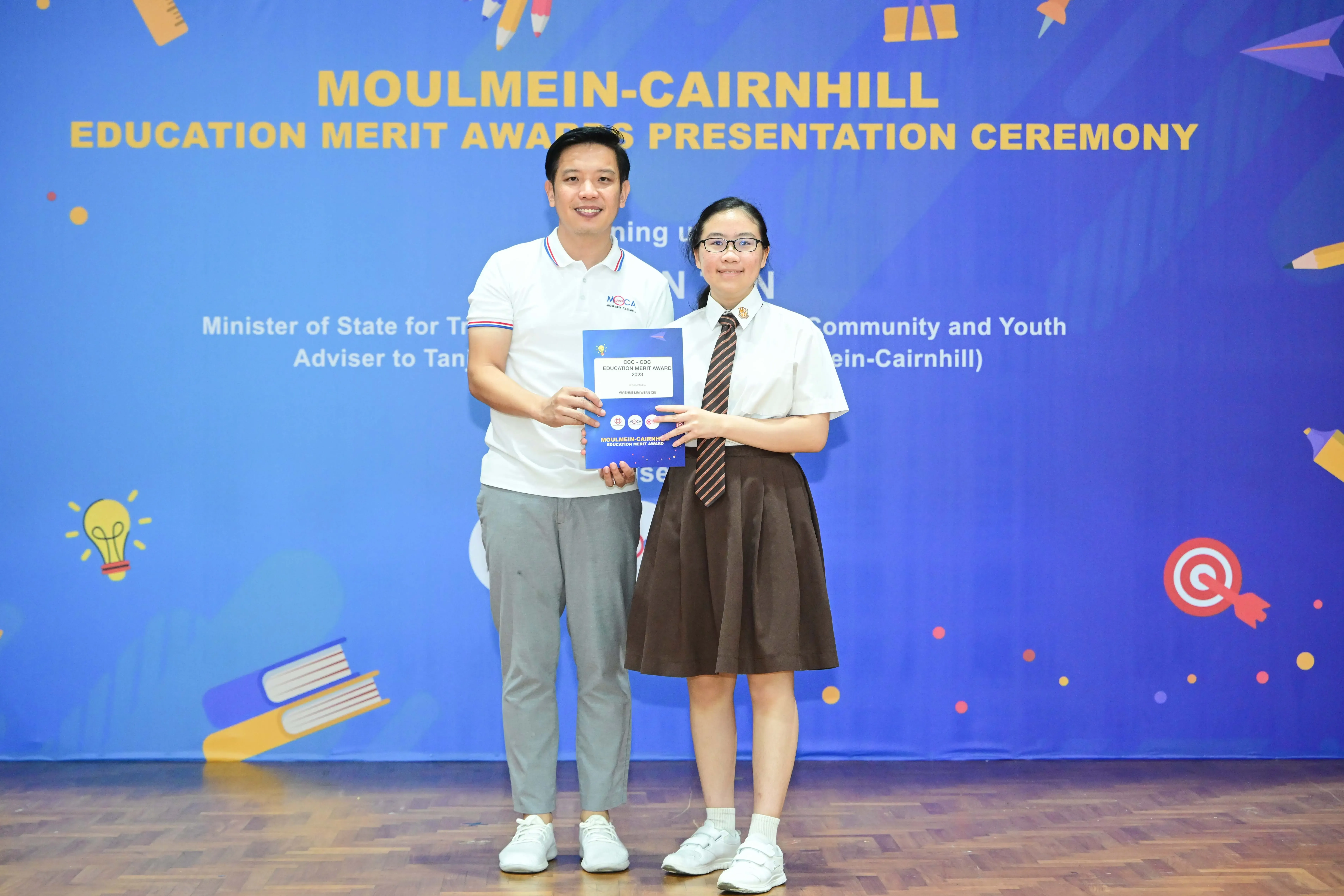 A man and a schoolgirl hold a certificate together at the Moulmein-Cairnhill Education Merit Awards Presentation Ceremony.