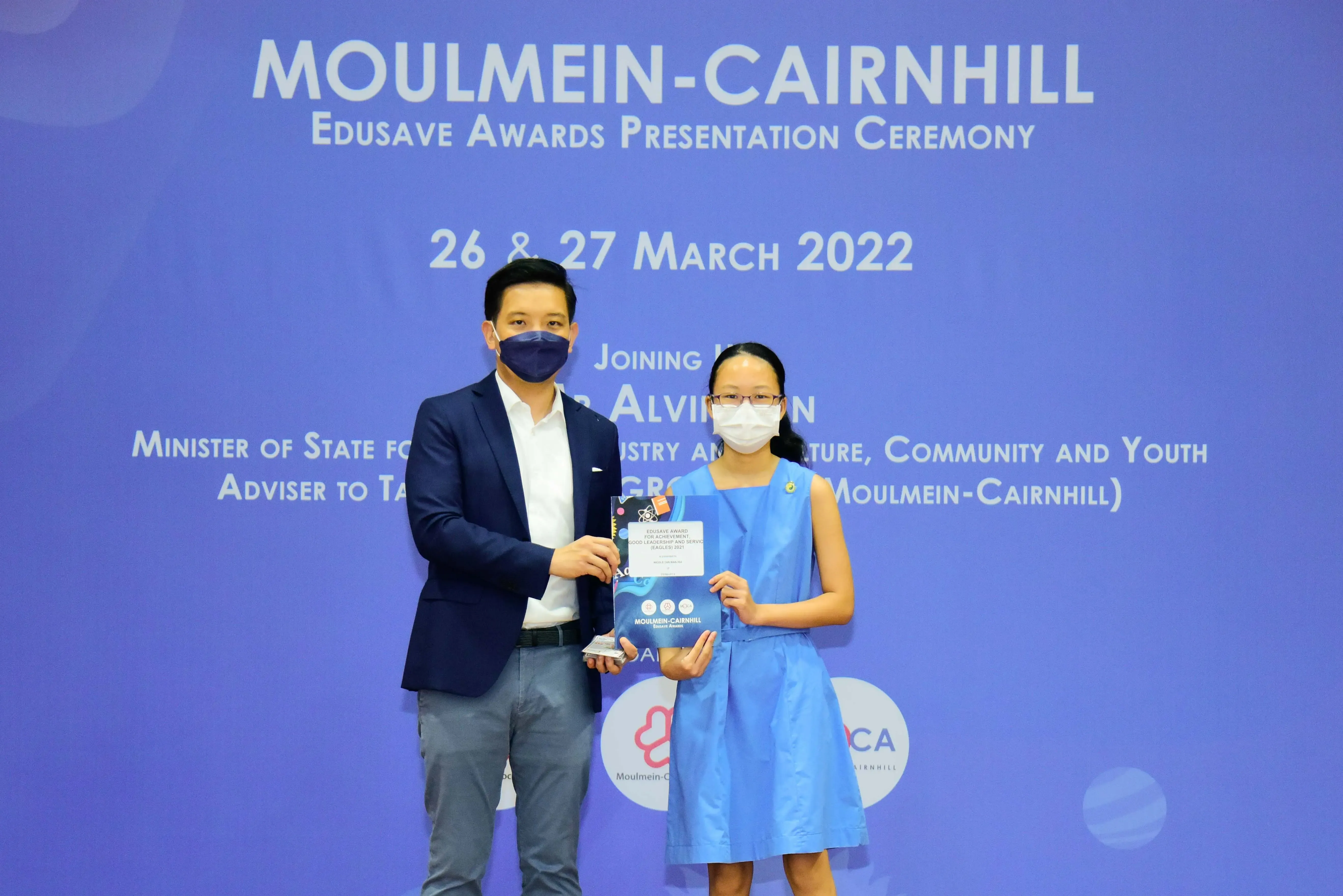 Two masked individuals hold an award at the Moulmein-Cairnhill Edusave Awards Presentation Ceremony, March 2022.